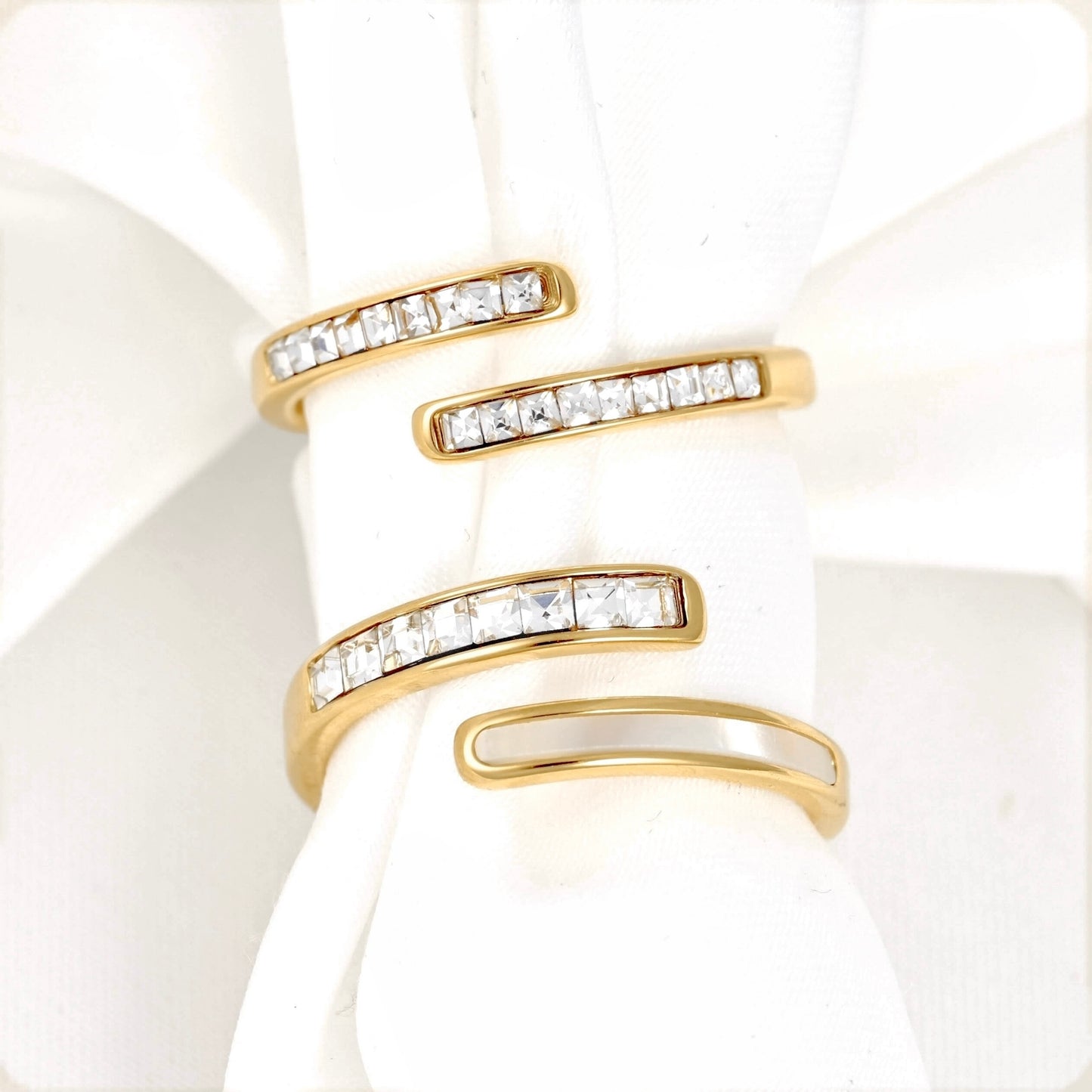 Two gold wrap-style rings with channel-set clear stones and shell inlay, displayed on a white fabric ring holder.