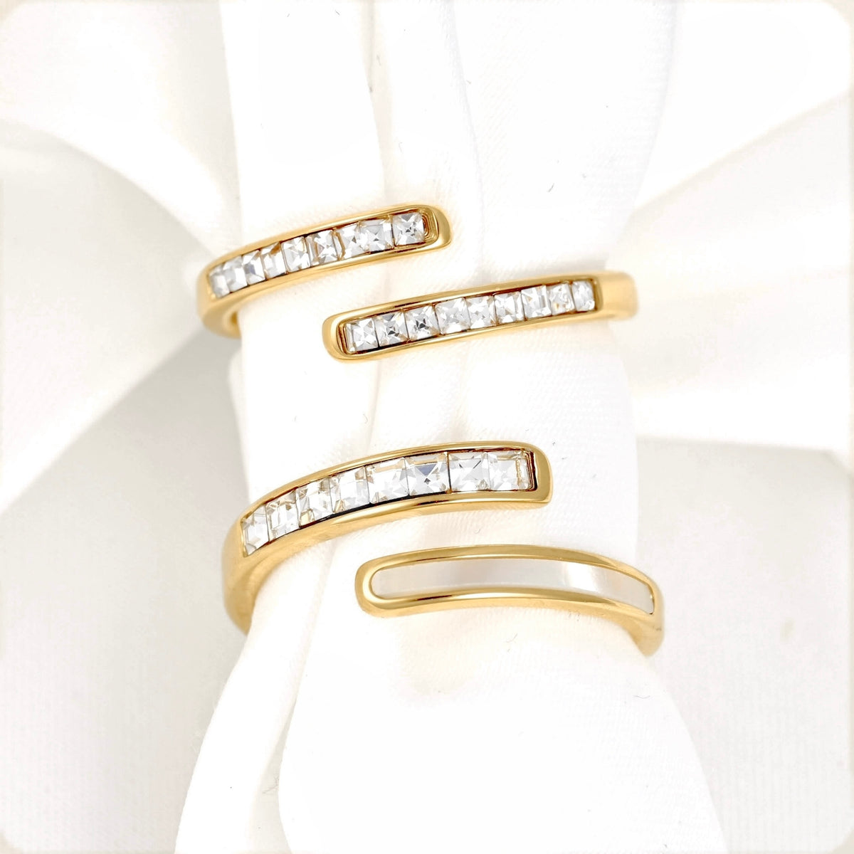 Two gold wrap-style rings with channel-set clear stones and shell inlay, displayed on a white fabric ring holder.