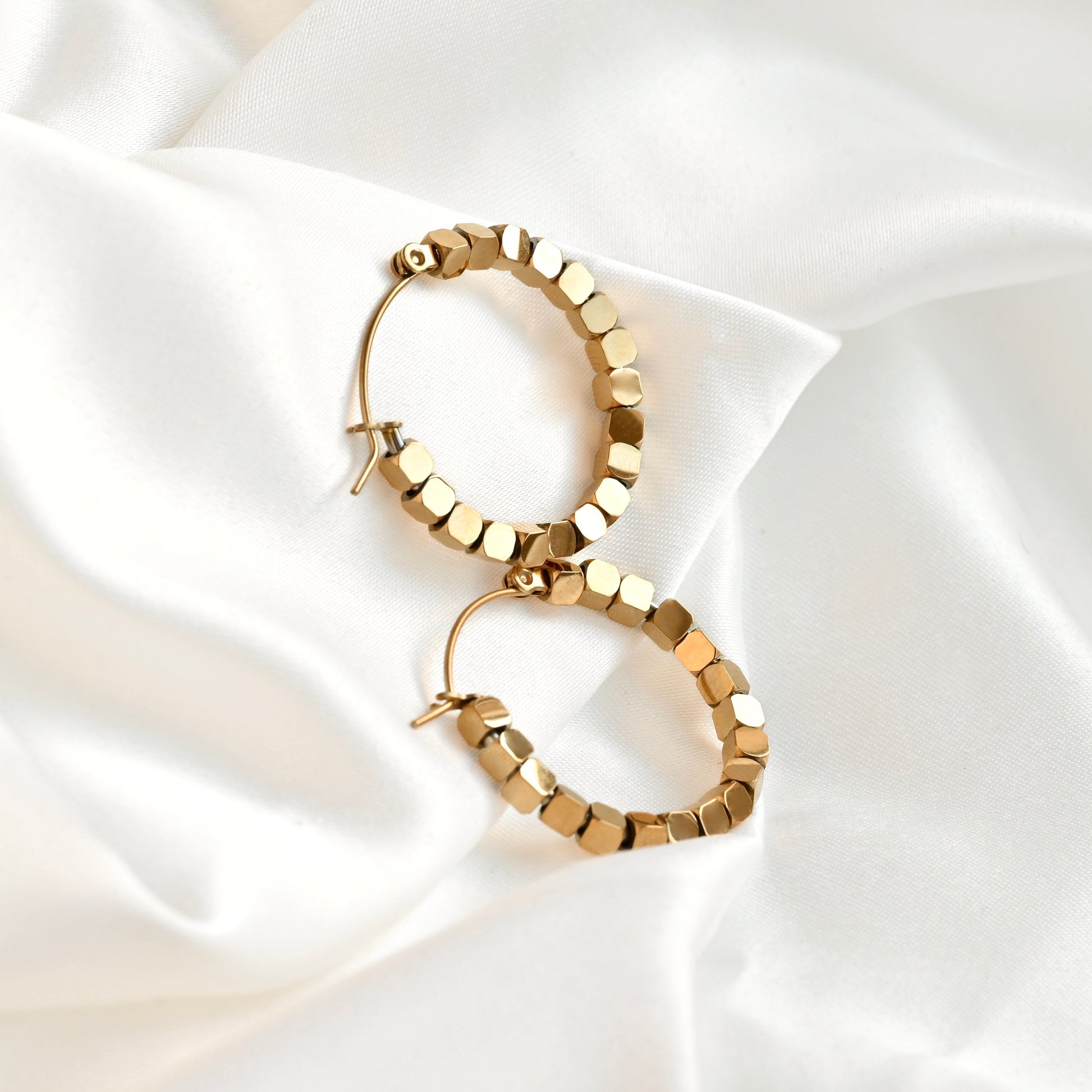 A pair of gold hoop earrings with a textured, faceted cube bead pattern, displayed on a draped white satin fabric background.