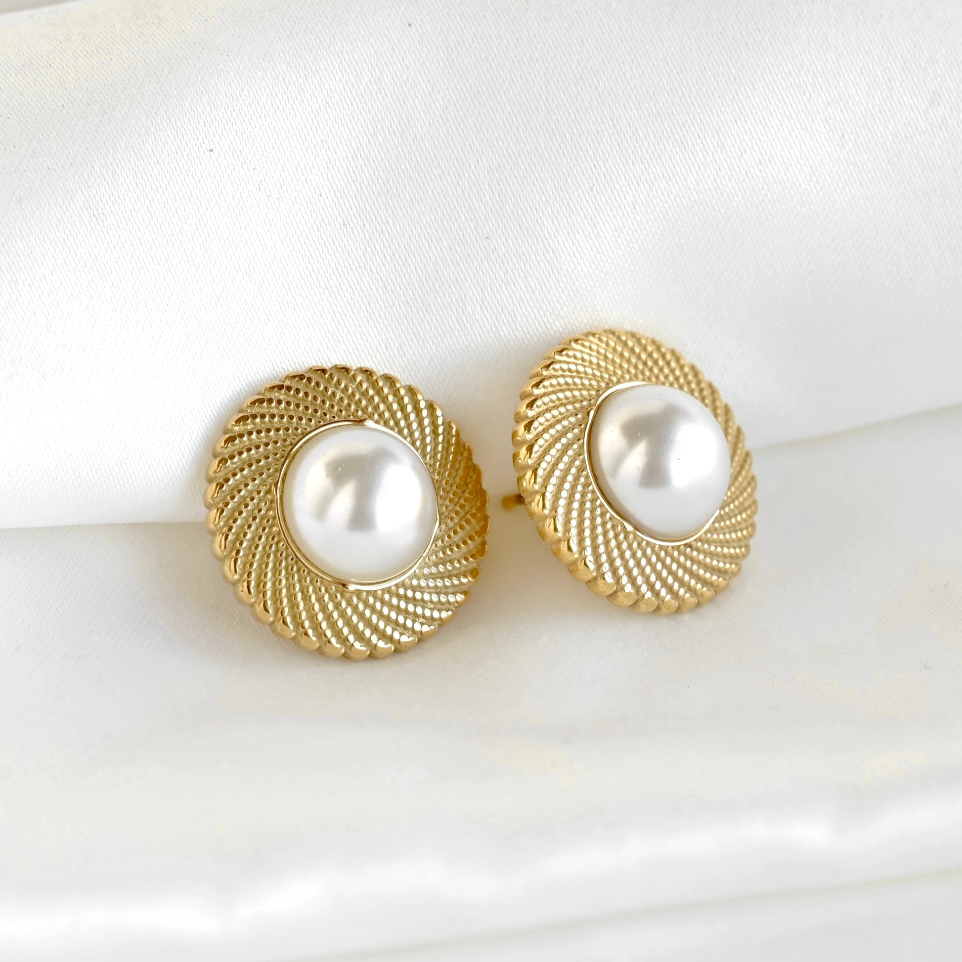A pair of gold round stud earrings with a textured rope-like border and a central white pearl, displayed on a soft white fabric background.