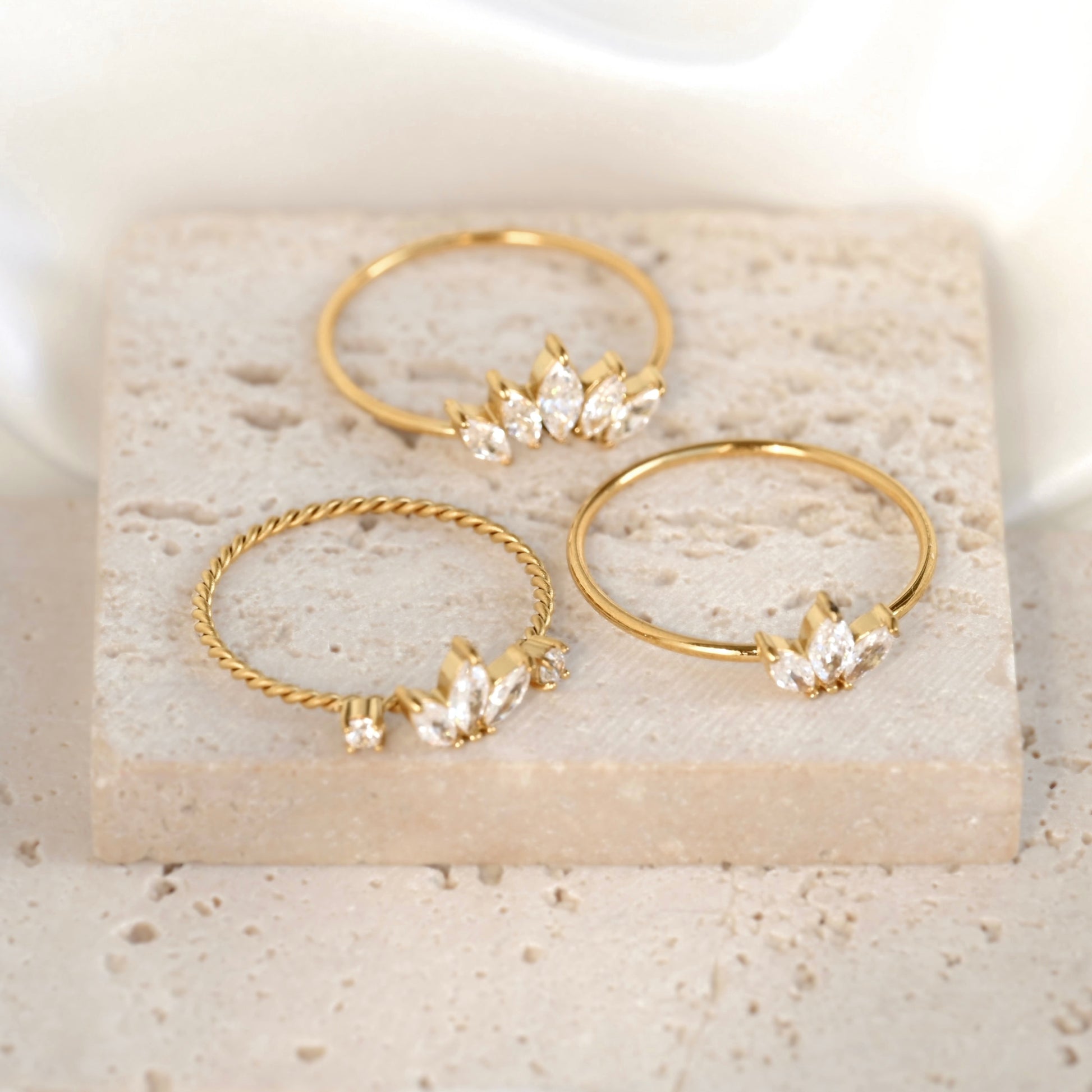 Three gold rings displayed on a light stone slab. Two rings are smooth bands with small clear crystal leaf clusters, and one ring is twisted with a matching crystal leaf cluster.