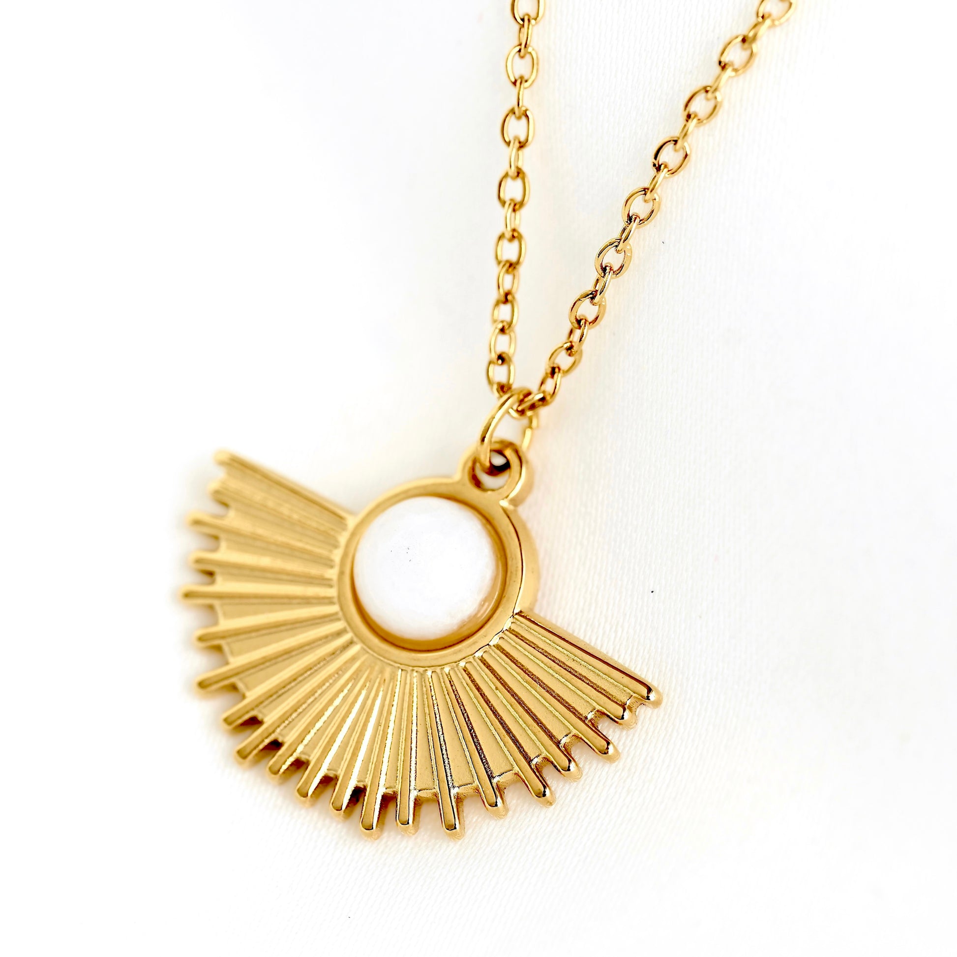 A gold pendant necklace featuring a sunburst fan design with a central white round stone, shown on a white textured fabric background.