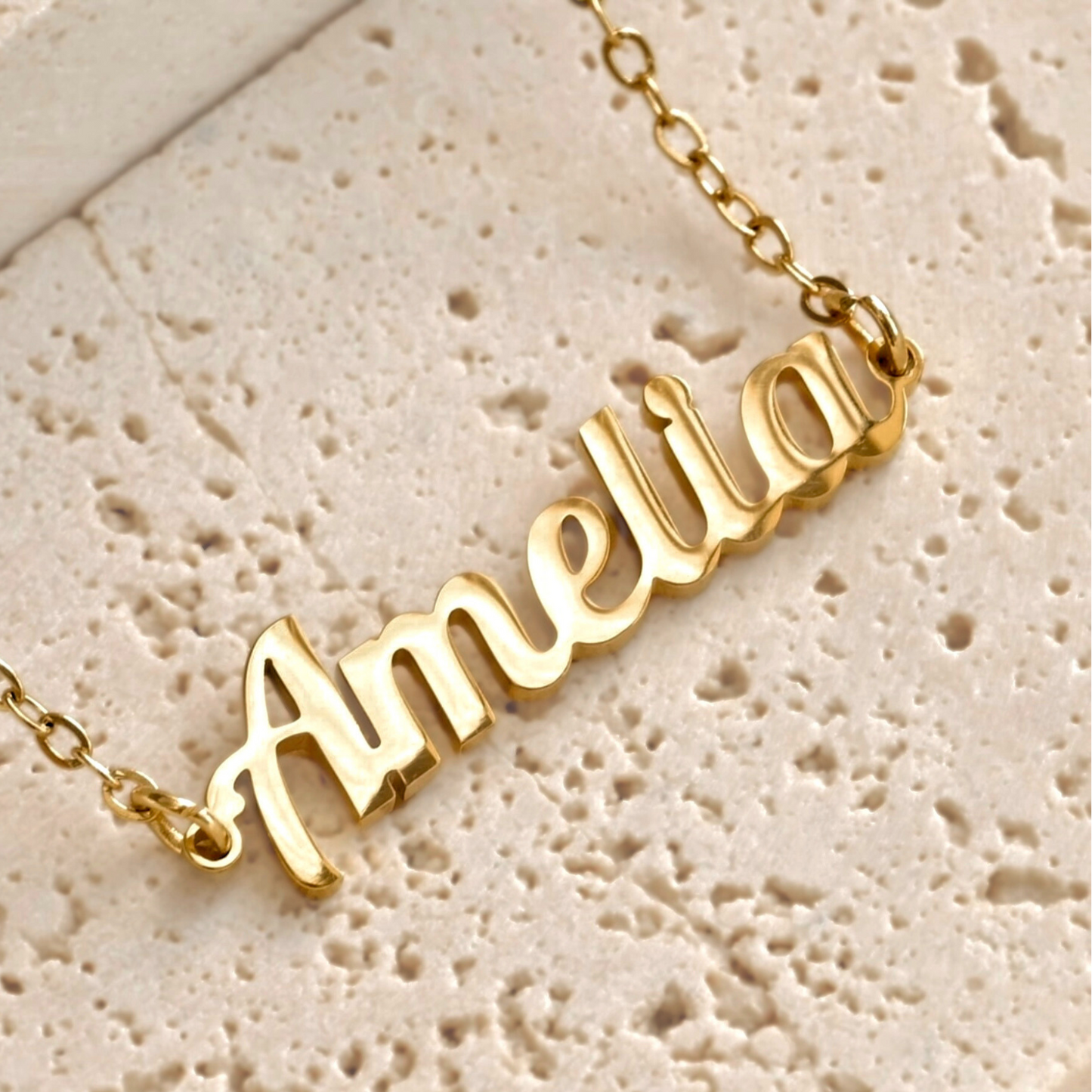 Gold script necklace with 'Amelia' pendant on a textured beige porous
 stone