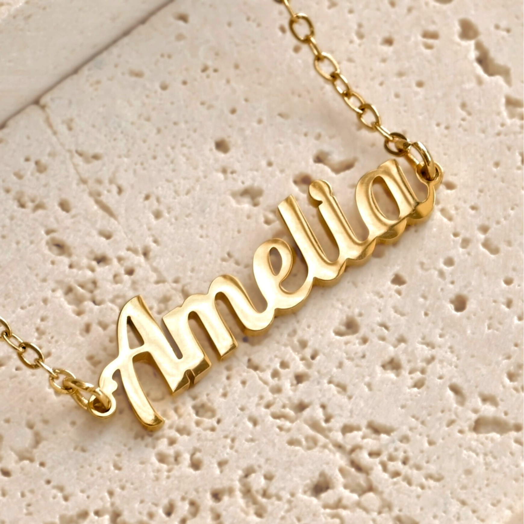 Gold script necklace with 'Amelia' pendant on a textured beige porous
 stone
