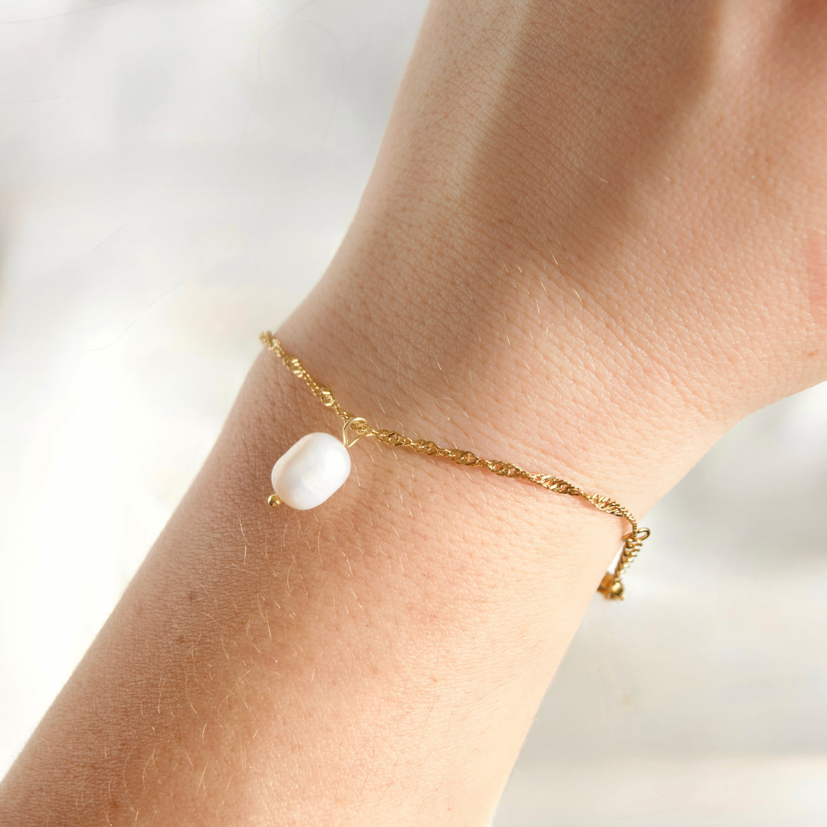 A delicate gold twisted chain bracelet with a single white baroque pearl charm, shown worn on a person's wrist against a soft, bright, out-of-focus background.