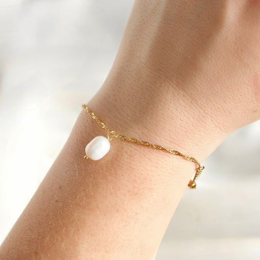 A delicate gold twisted chain bracelet with a single white baroque pearl charm, shown worn on a person's wrist against a soft, bright, out-of-focus background.