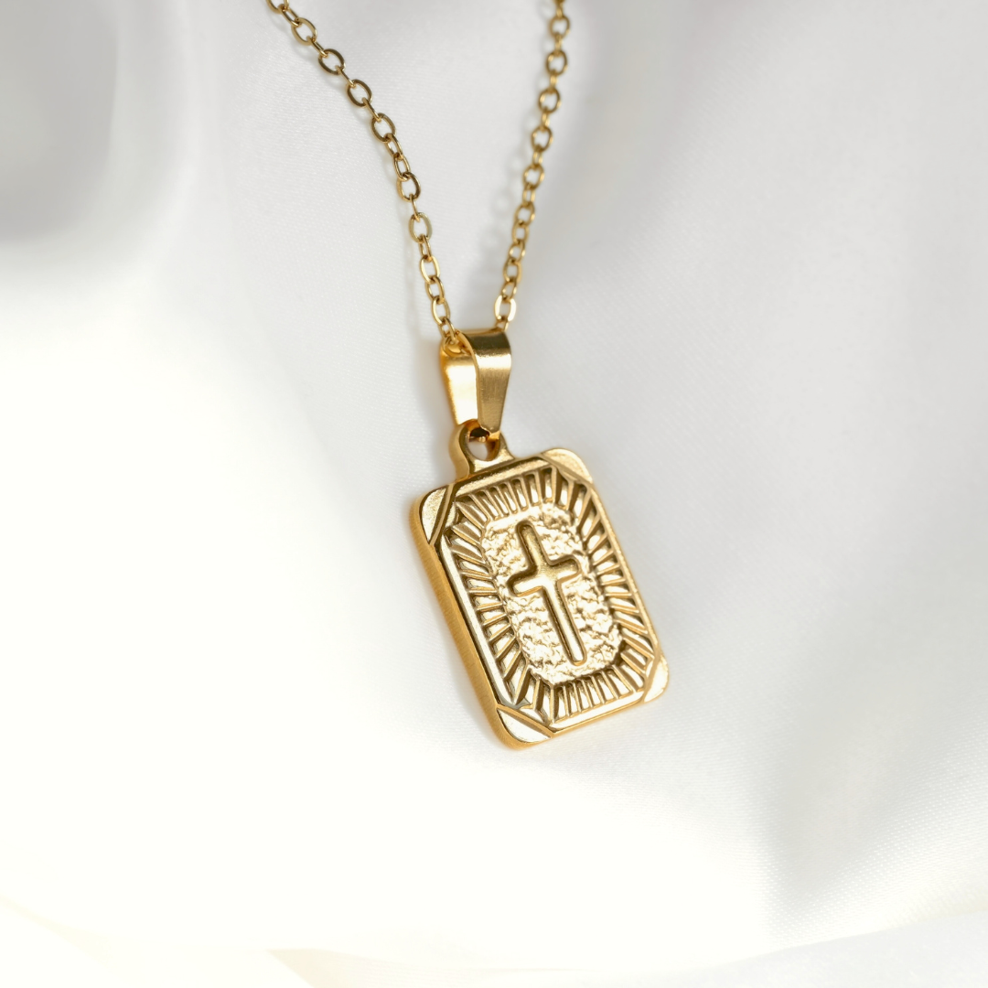 A gold-toned religious pendant necklace featuring a rectangular engraved cross on a chain, displayed on a soft white fabric background.