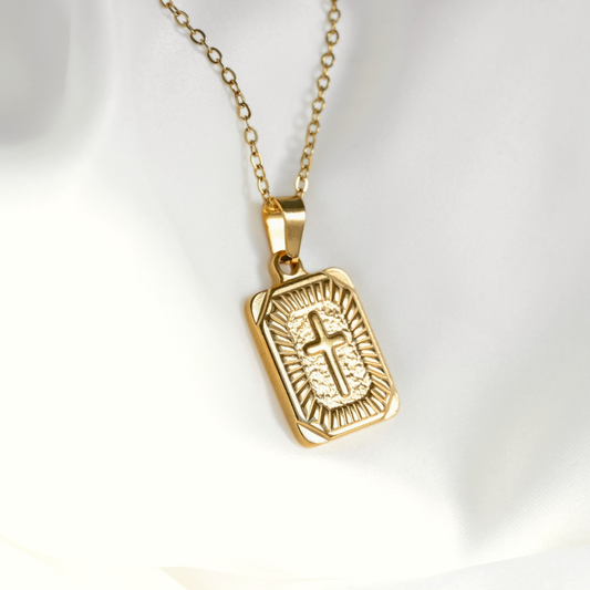 A gold-toned religious pendant necklace featuring a rectangular engraved cross on a chain, displayed on a soft white fabric background.