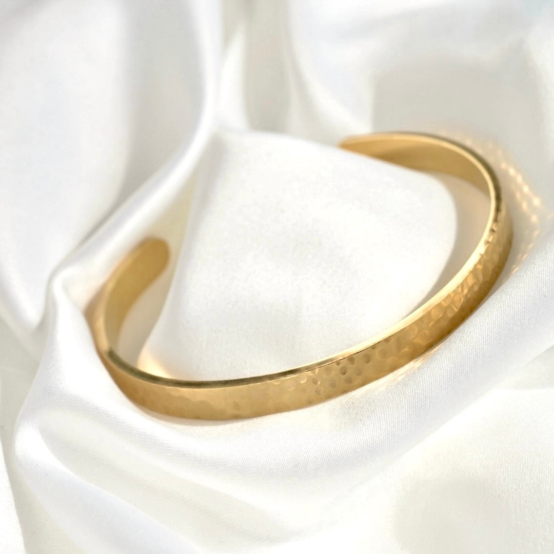 A hammered gold-tone open cuff bangle bracelet displayed on a soft white satin fabric background.