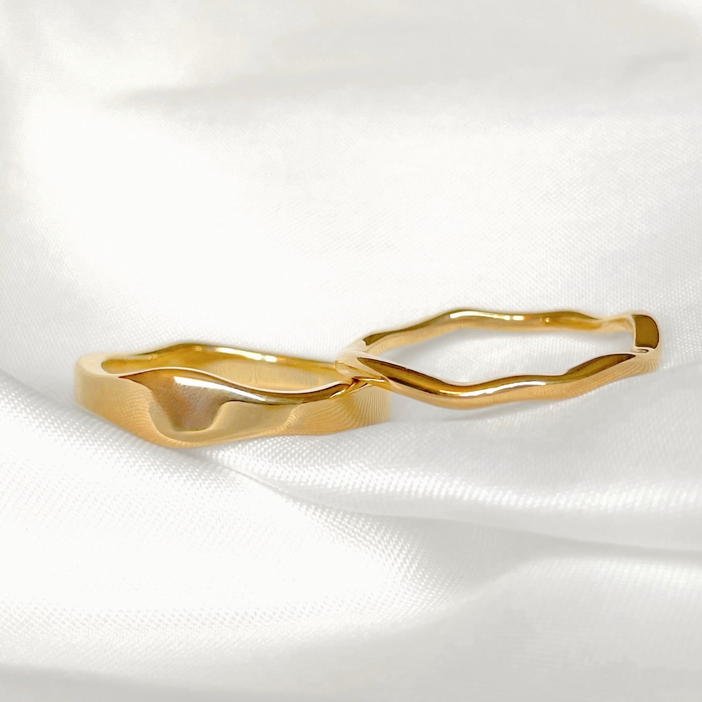 Molten gold rings with wavy design, one thick and one thin, on a white satin background