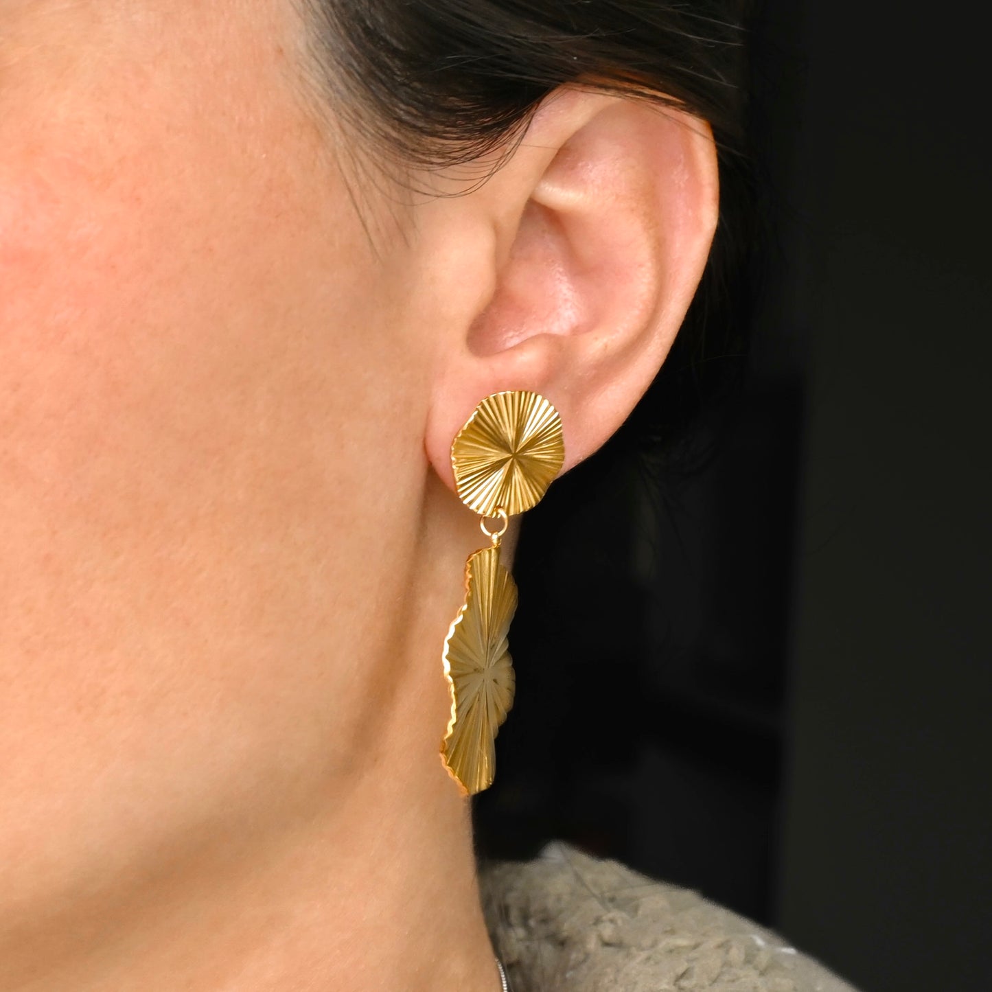 Close-up of a person wearing gold sunburst drop earrings with a dark background