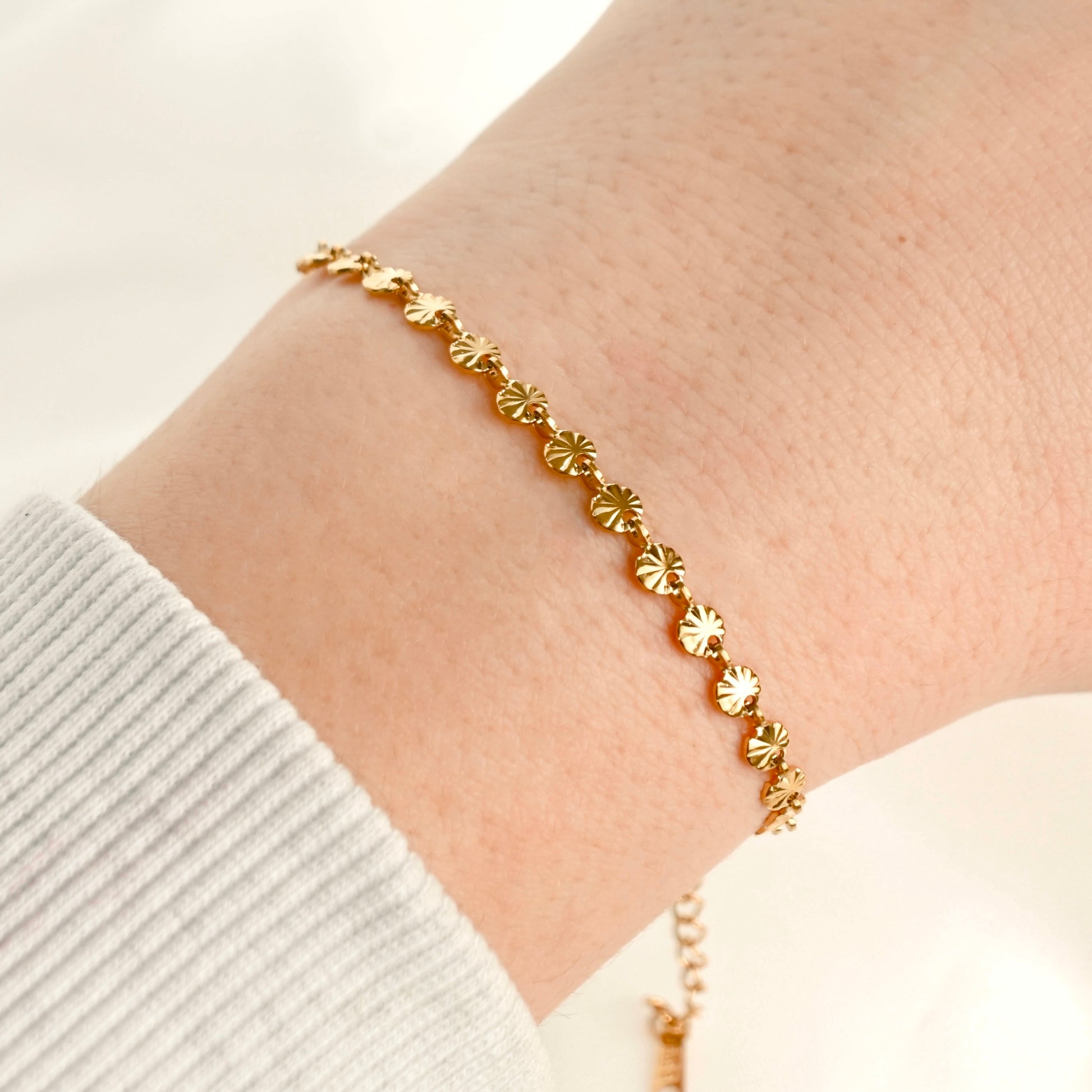 A delicate gold chain bracelet with small, textured sunburst charms, shown worn on a person's wrist over a white ribbed knit sleeve.