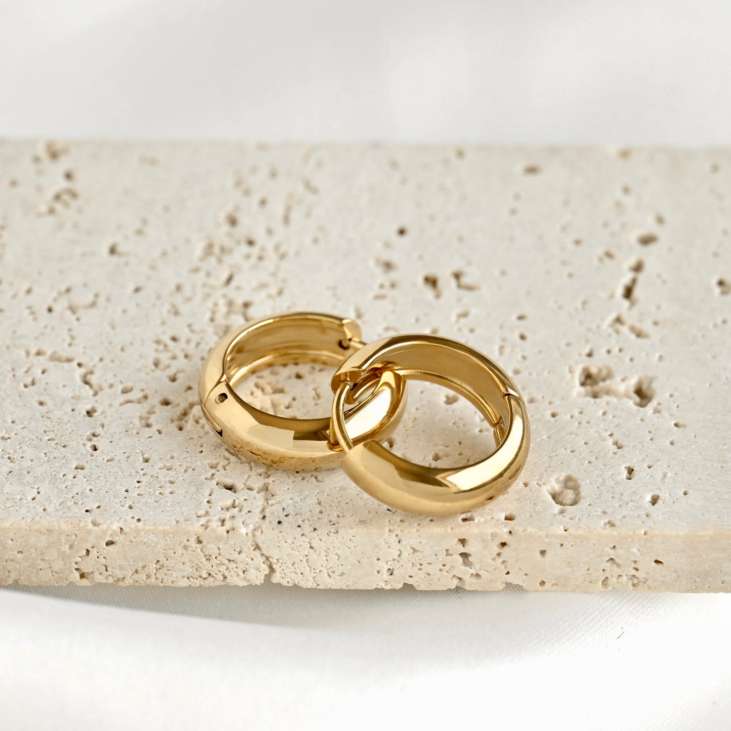 Two polished bold gold huggie hoop earrings resting on a textured beige stone slab against a clean white background.