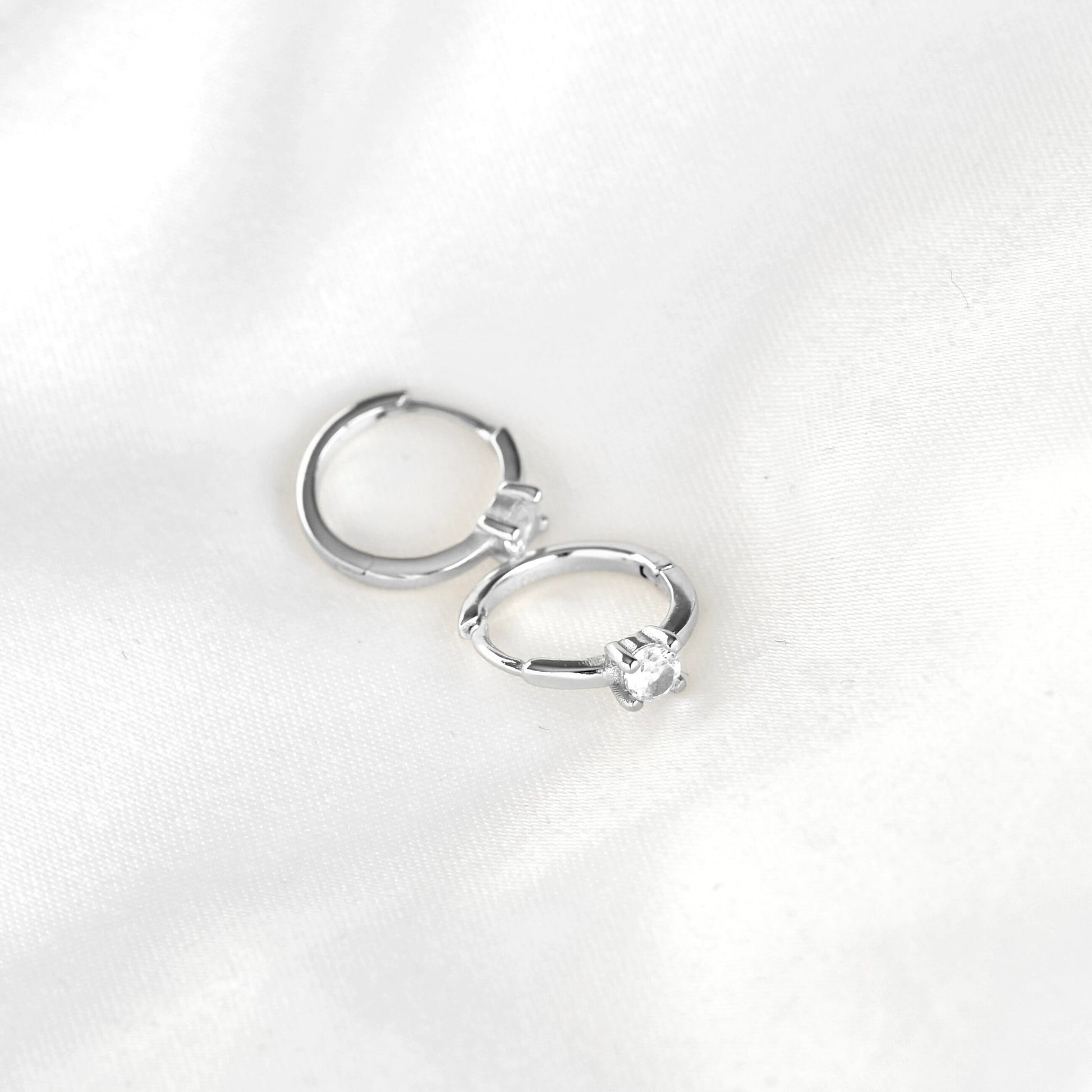 A pair of small silver hoop earrings with a single clear gemstone in each hoop, displayed on a white textured fabric background.