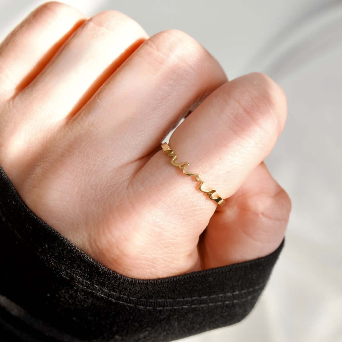 A close-up of a hand wearing a delicate gold ring with a wavy, scalloped edge design, set against a soft, light gray background.