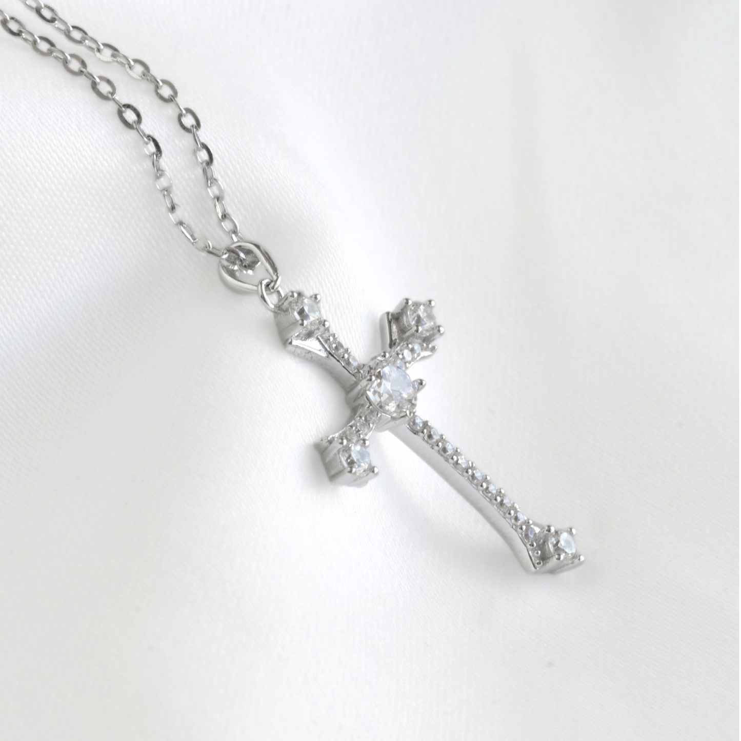 A silver cross pendant necklace adorned with clear crystals, resting on a white satin fabric background.