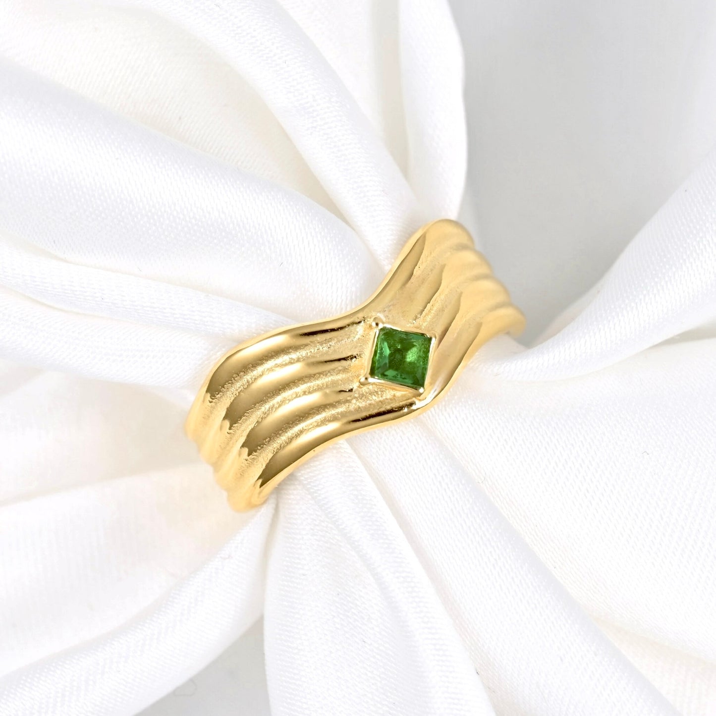 A gold ring with a textured, wavy band and a small square green gemstone, displayed on a white fabric ring holder.