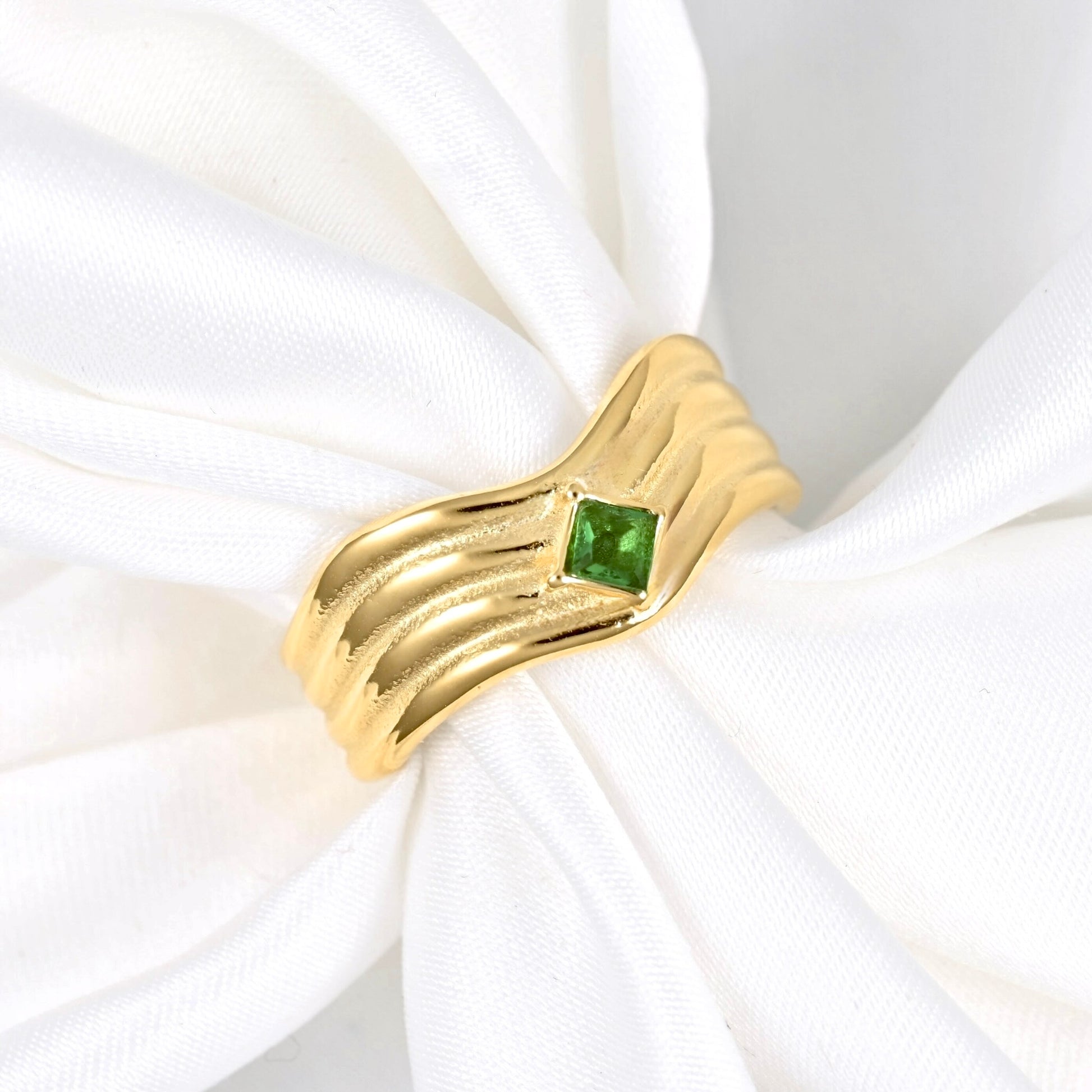 A gold ring with a textured, wavy band and a small square green gemstone, displayed on a white fabric ring holder.