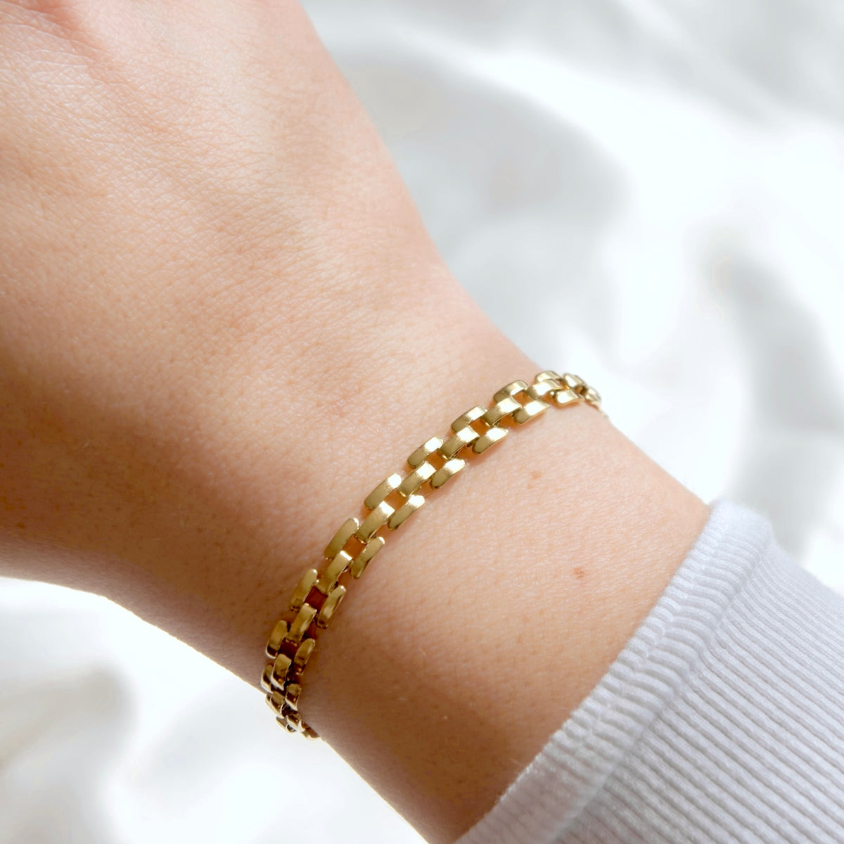 A close-up of a gold chain bracelet worn on a person's wrist, with a white ribbed cuff visible at the side against a soft white fabric background.