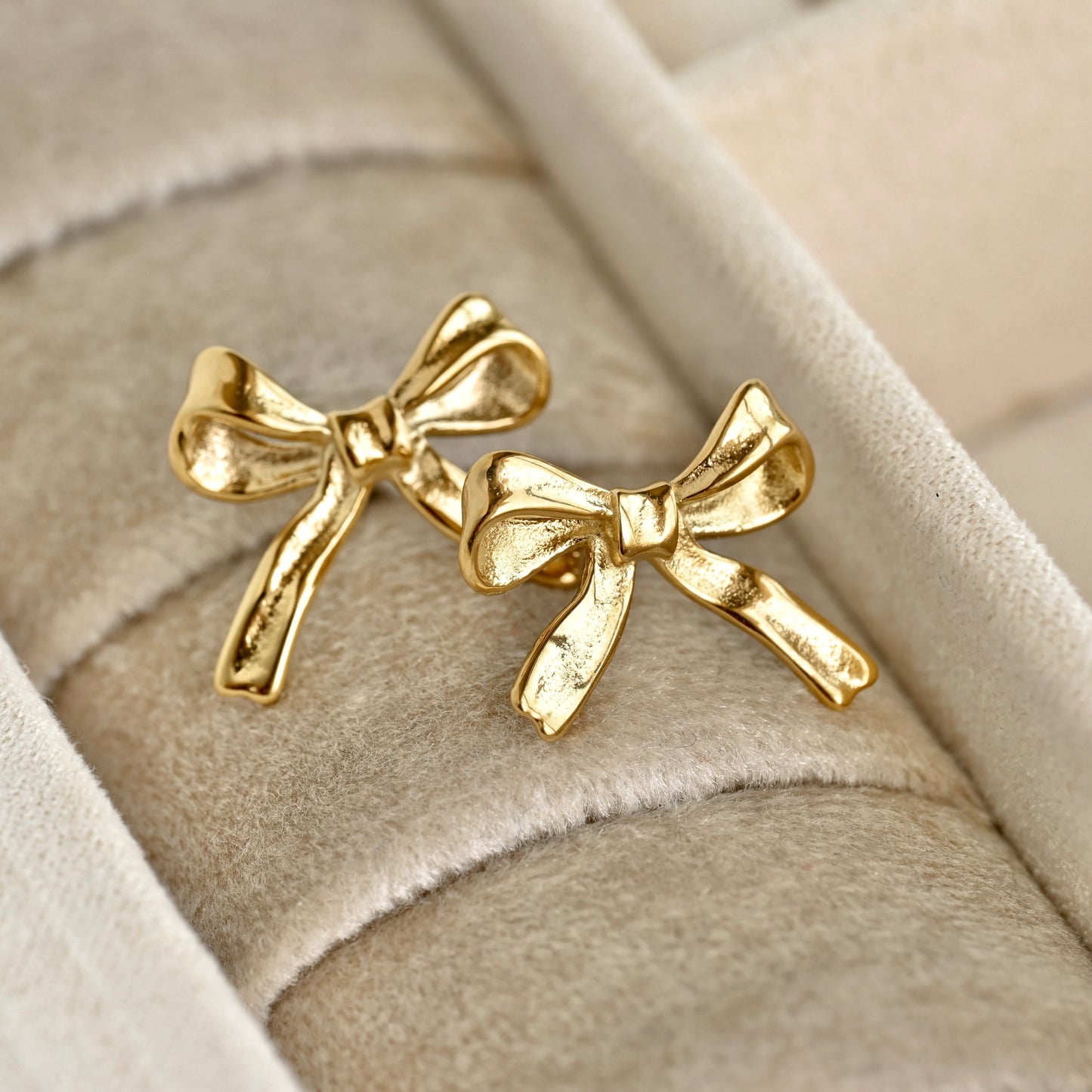 A pair of gold bow tie stud earrings displayed on a beige velvet jewelry ring roll.