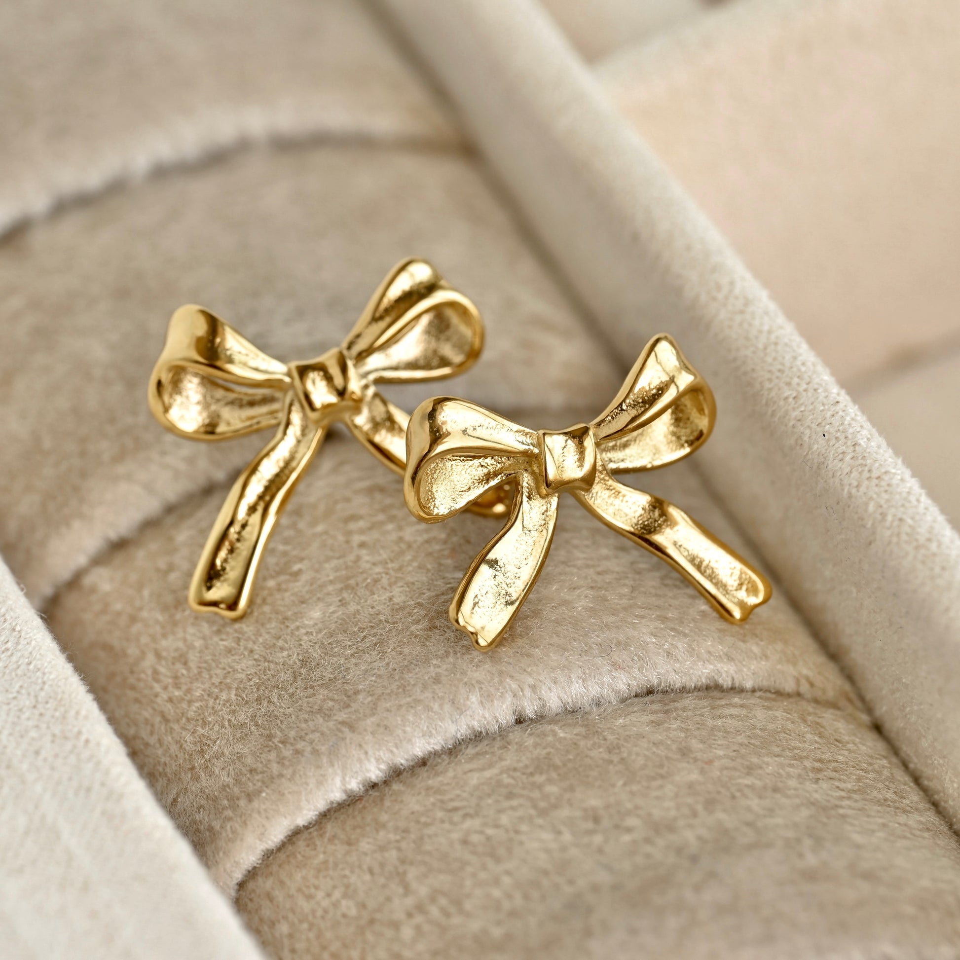 A pair of gold bow tie stud earrings displayed on a beige velvet jewelry ring roll.