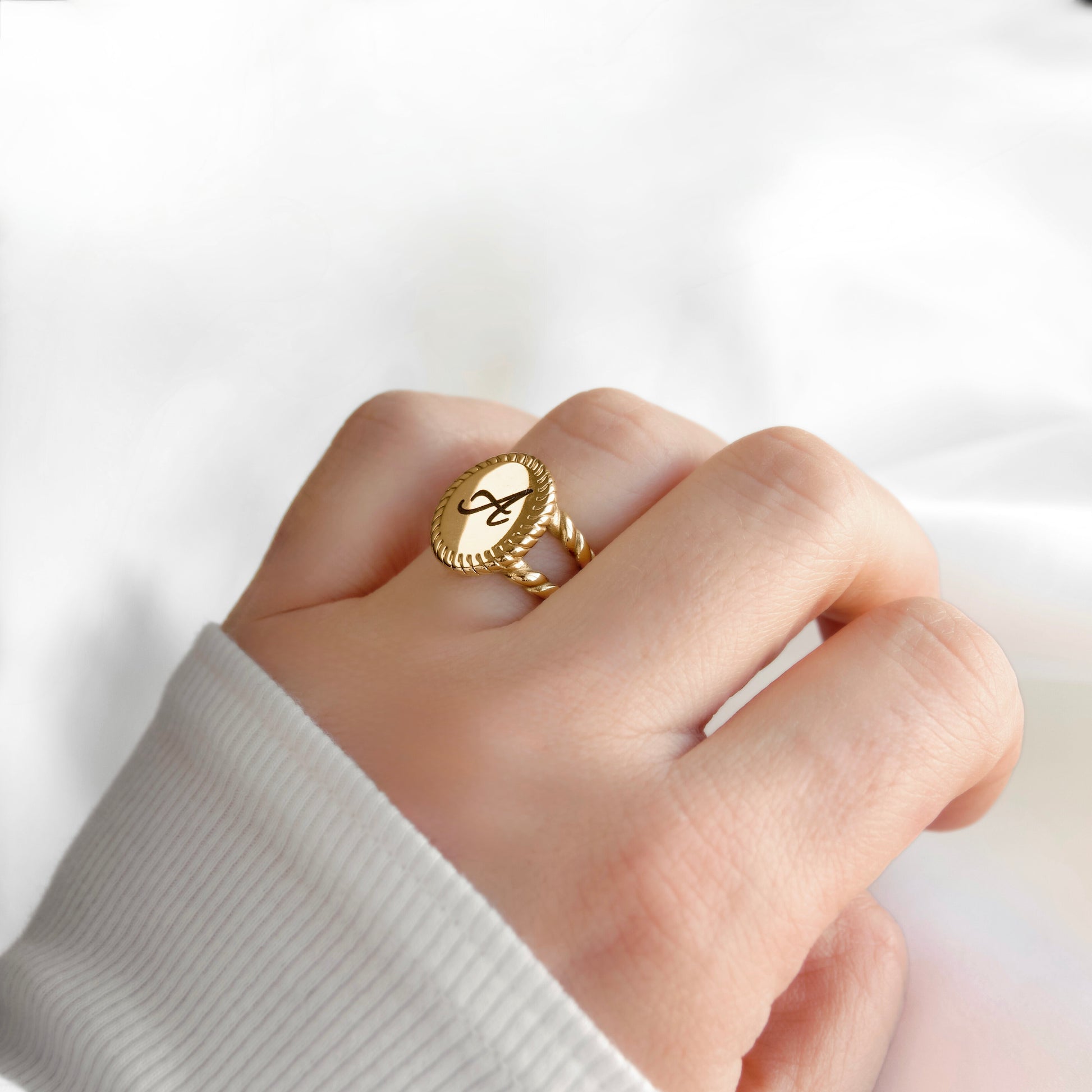 A close-up of a hand wearing a gold signet-style ring with a circular face and script lettering, against a soft white background.