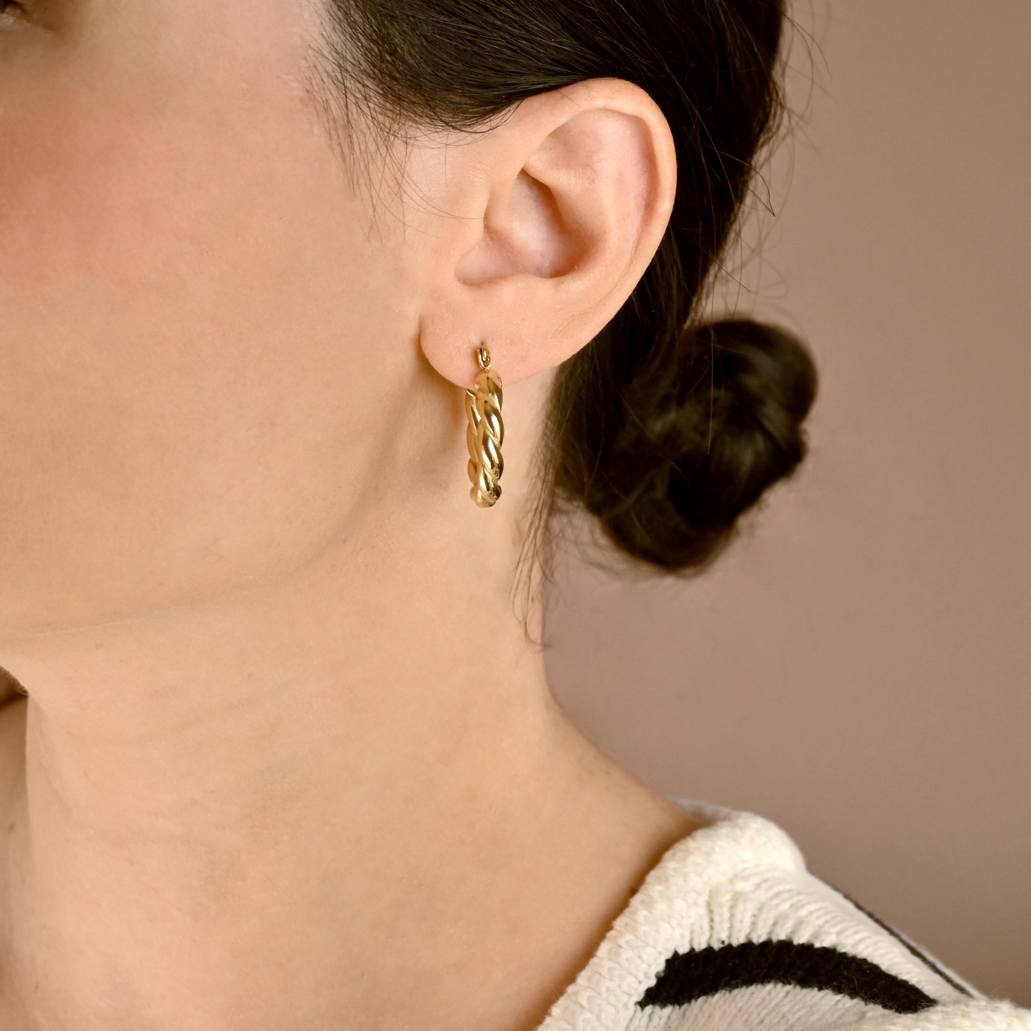 A close-up of a woman's ear wearing a small twisted gold hoop earring, with her hair tied back and a neutral beige background.