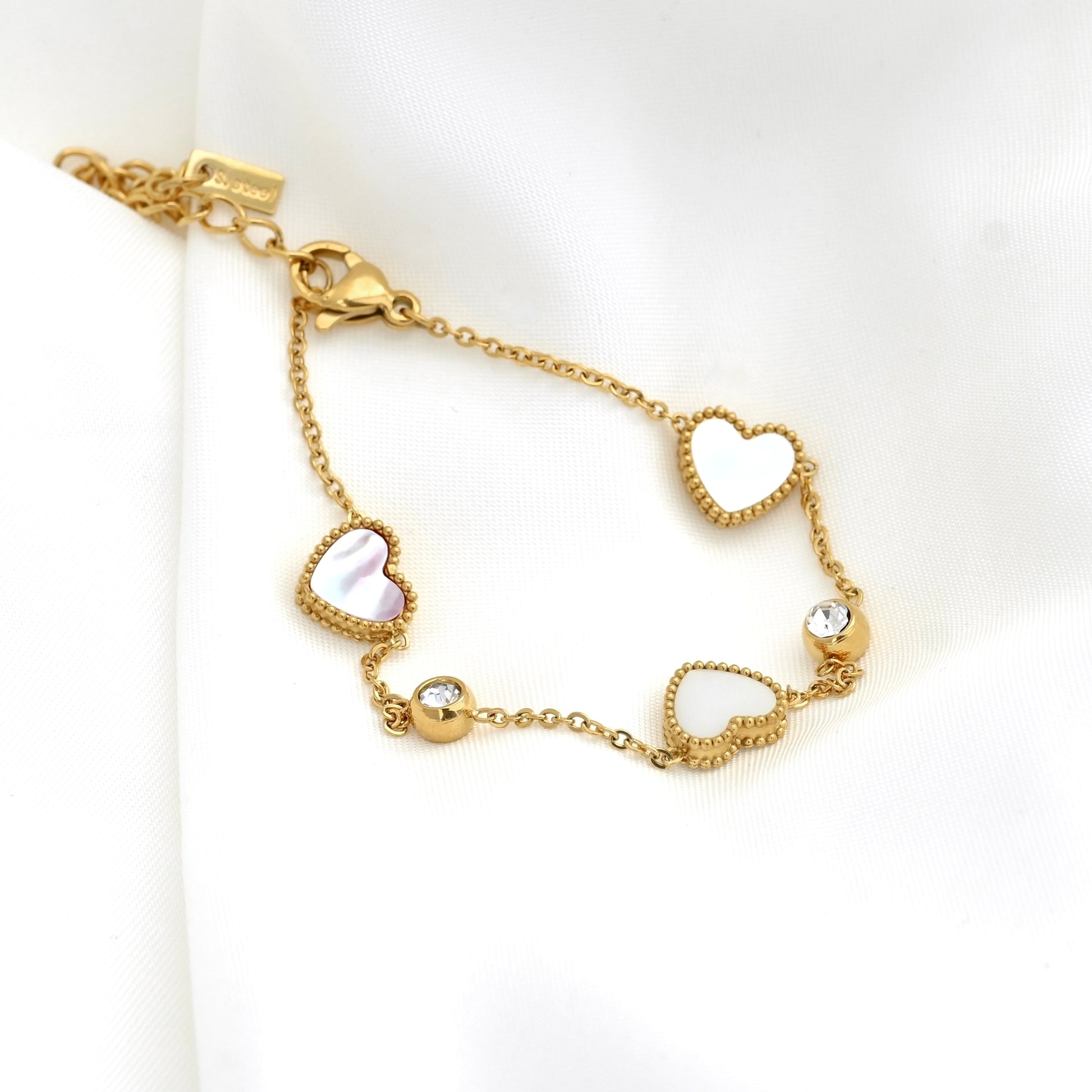 A gold chain bracelet featuring three  white mother-of-pearl hearts with milgrain details, and two small round clear CZ accents, displayed on a white fabric background.