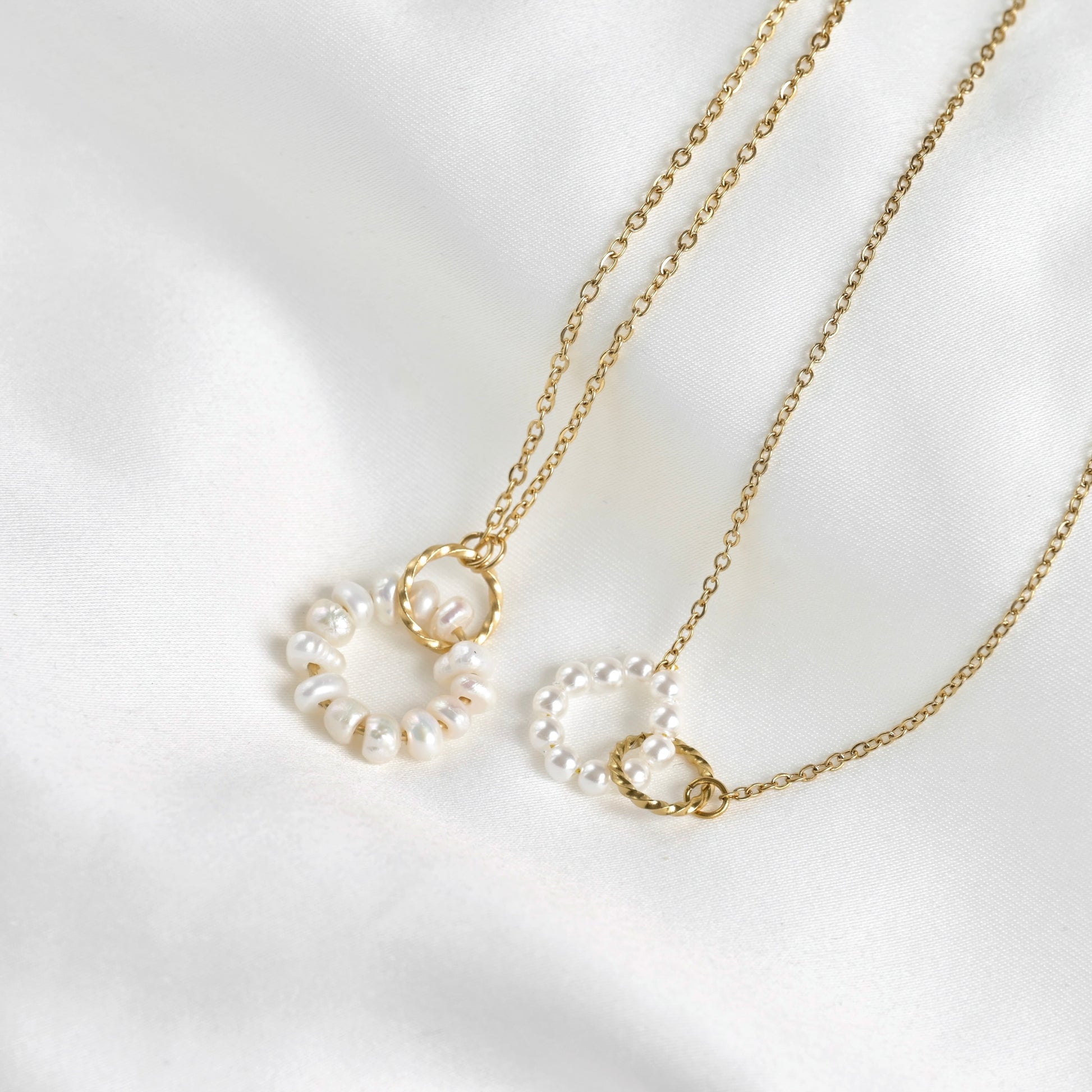 Two gold chain necklaces with circular pearl bead pendants, displayed on a soft white satin fabric background.