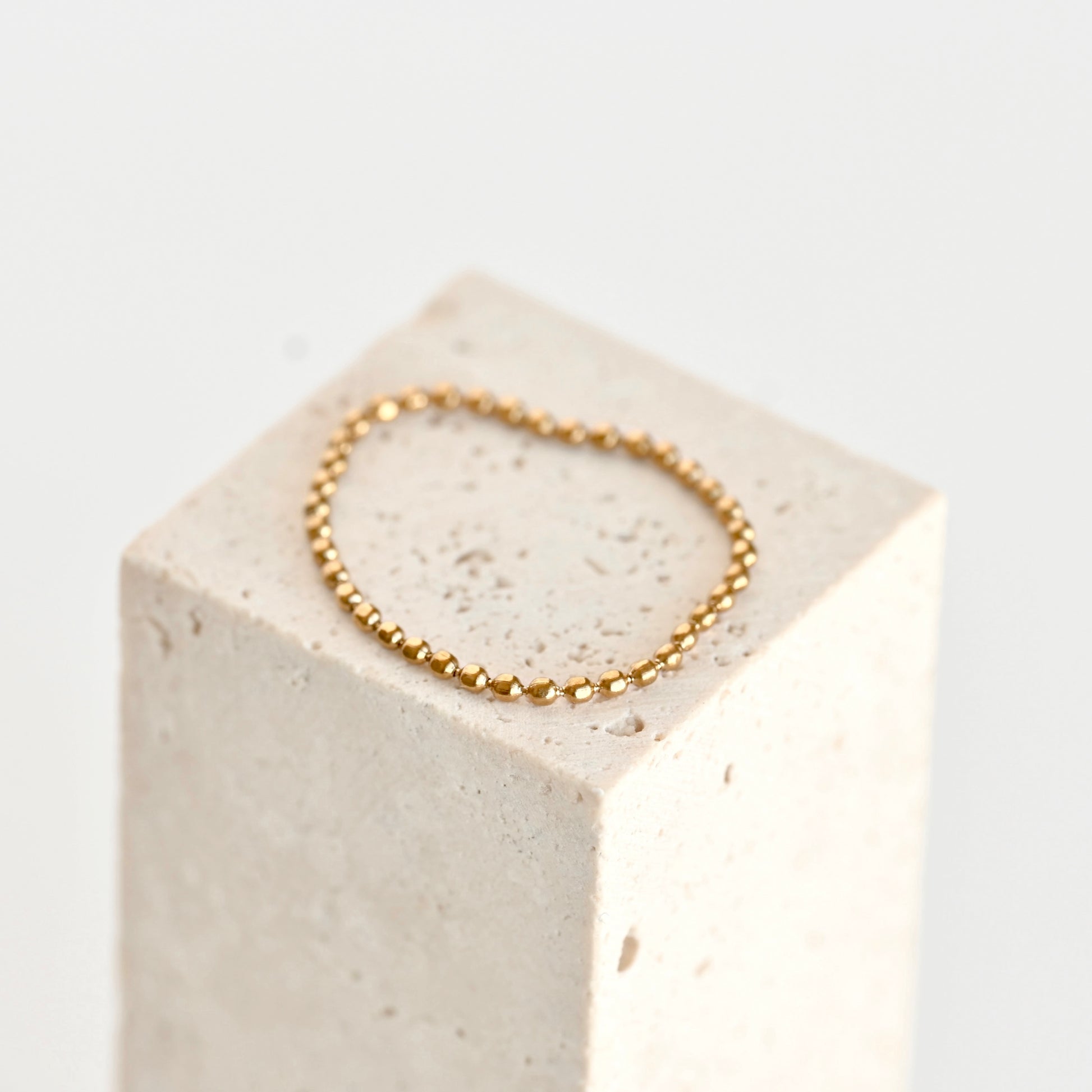 A delicate gold beaded chain ring displayed on a textured light beige stone block against a clean white background.