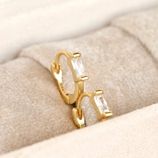 A pair of gold huggie hoop earrings with rectangular clear baguette-cut stones, displayed on a beige textured velvet jewelry stand.
