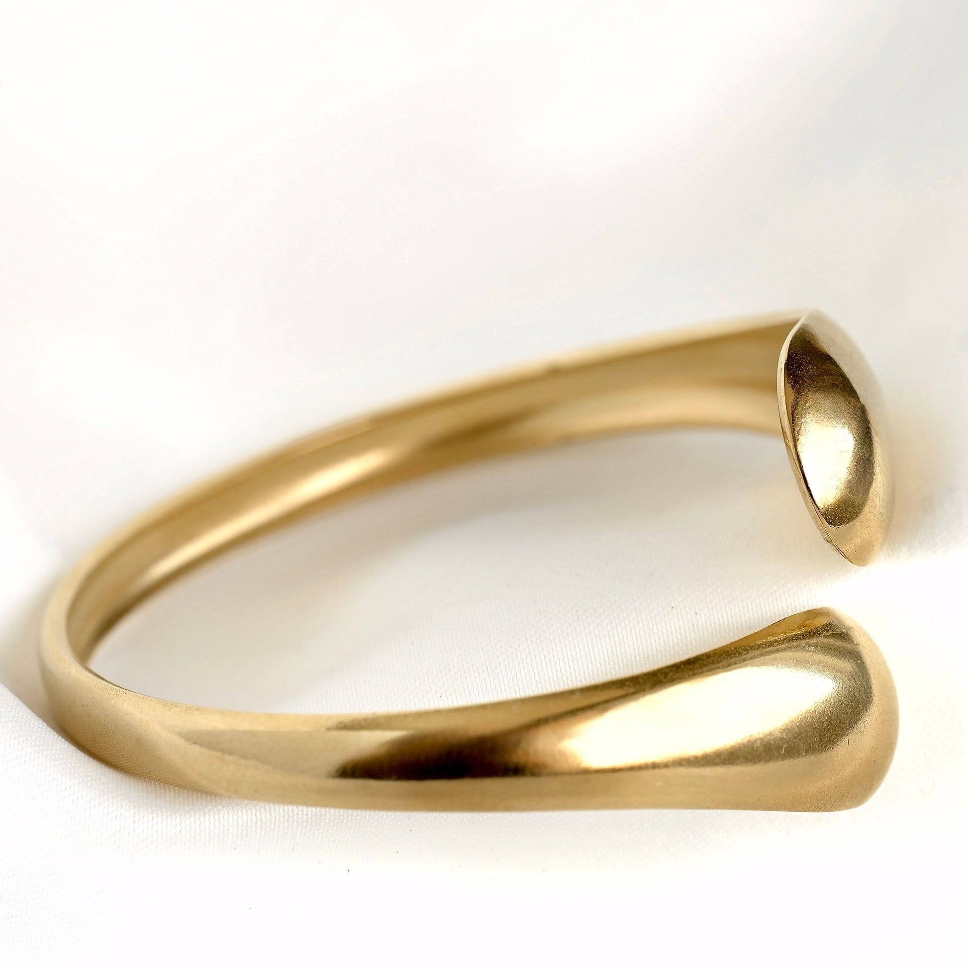 A polished gold-tone open cuff ring, also known as a bypass ring, displayed on a clean white textured fabric background.