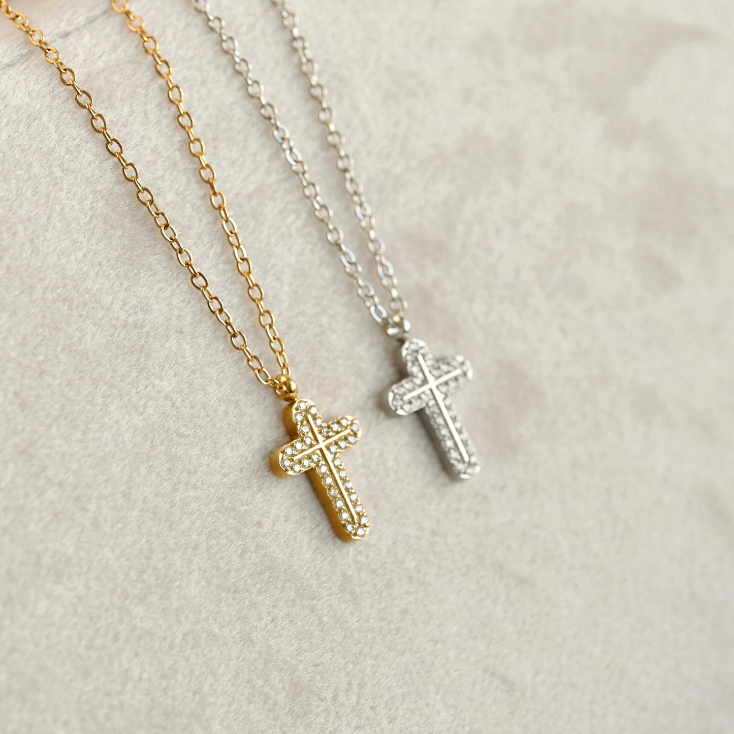 Two cross necklaces, one gold and one silver, both adorned with small clear stones, displayed on a textured light beige surface.