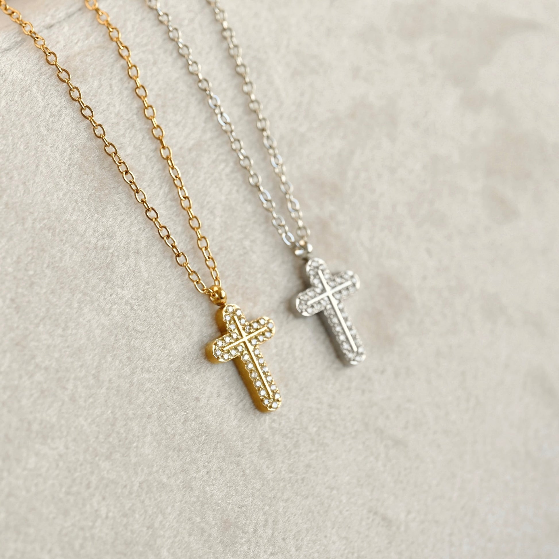 Two cross necklaces, one gold and one silver, both adorned with small clear stones, displayed on a textured light beige surface.