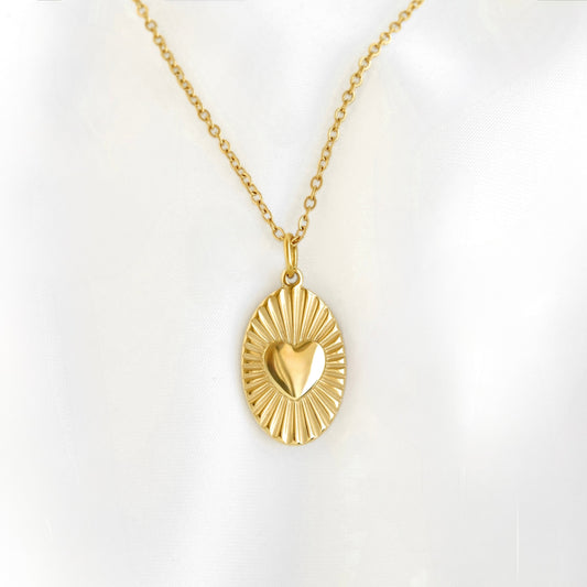 A gold pendant necklace featuring a sunburst oval design with a polished heart at the center, displayed on a soft white background.
