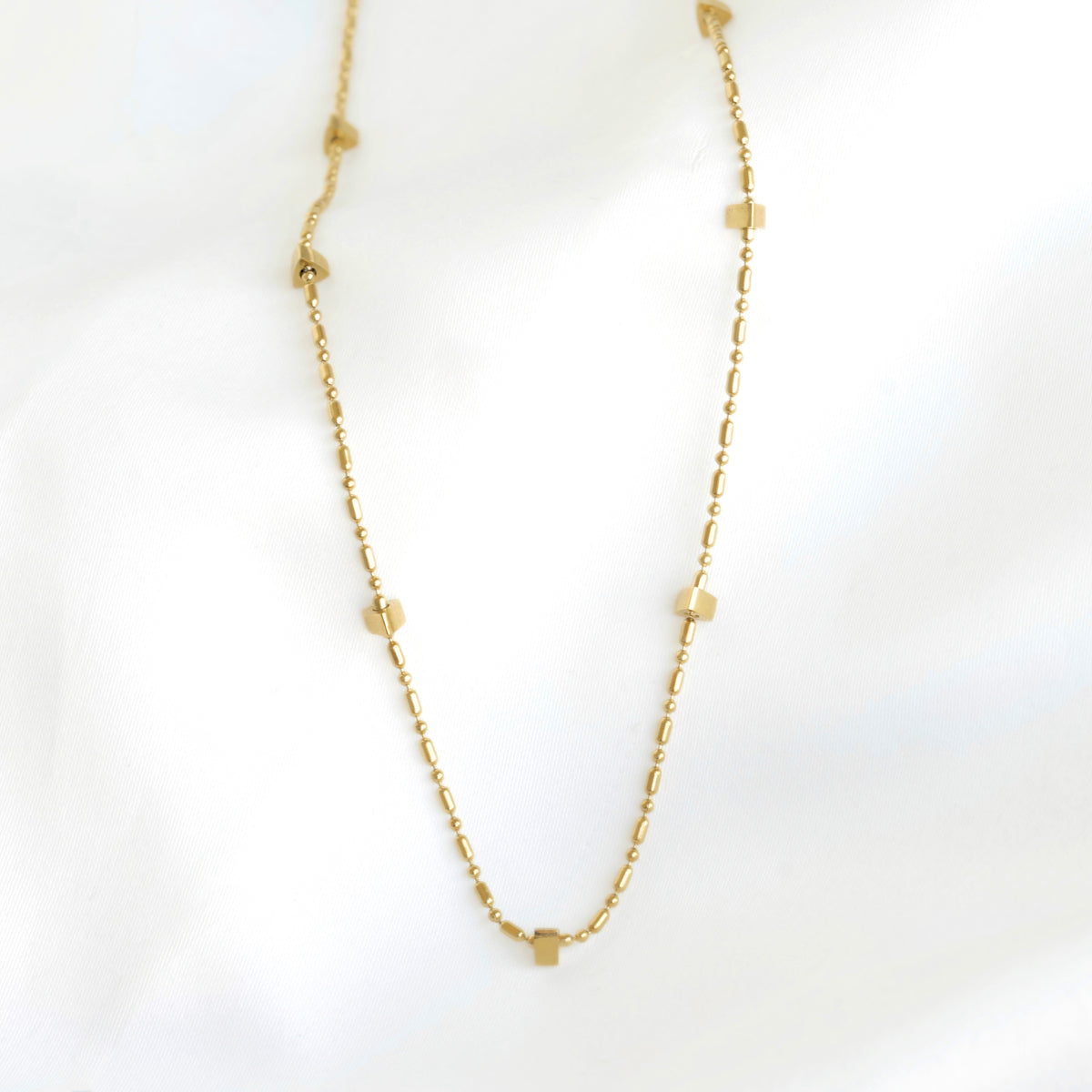 A delicate gold chain necklace featuring small, evenly spaced geometric charms, displayed on a soft white fabric background.