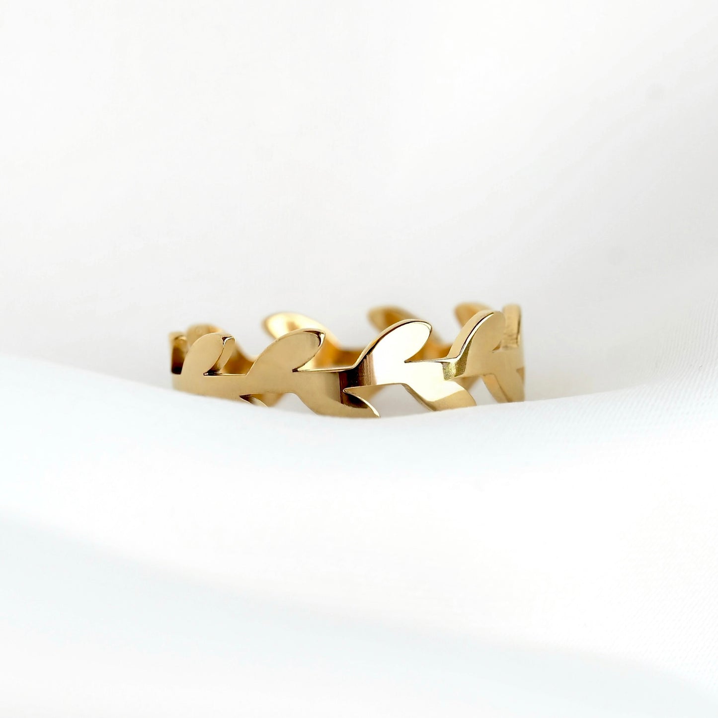 A gold-toned olive branch ring with a continuous leaf or vine motif, resting on a soft white fabric surface.