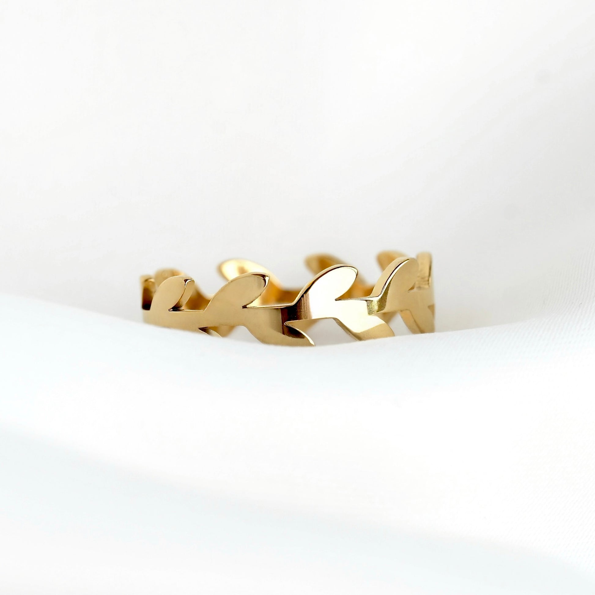 A gold-toned olive branch ring with a continuous leaf or vine motif, resting on a soft white fabric surface.