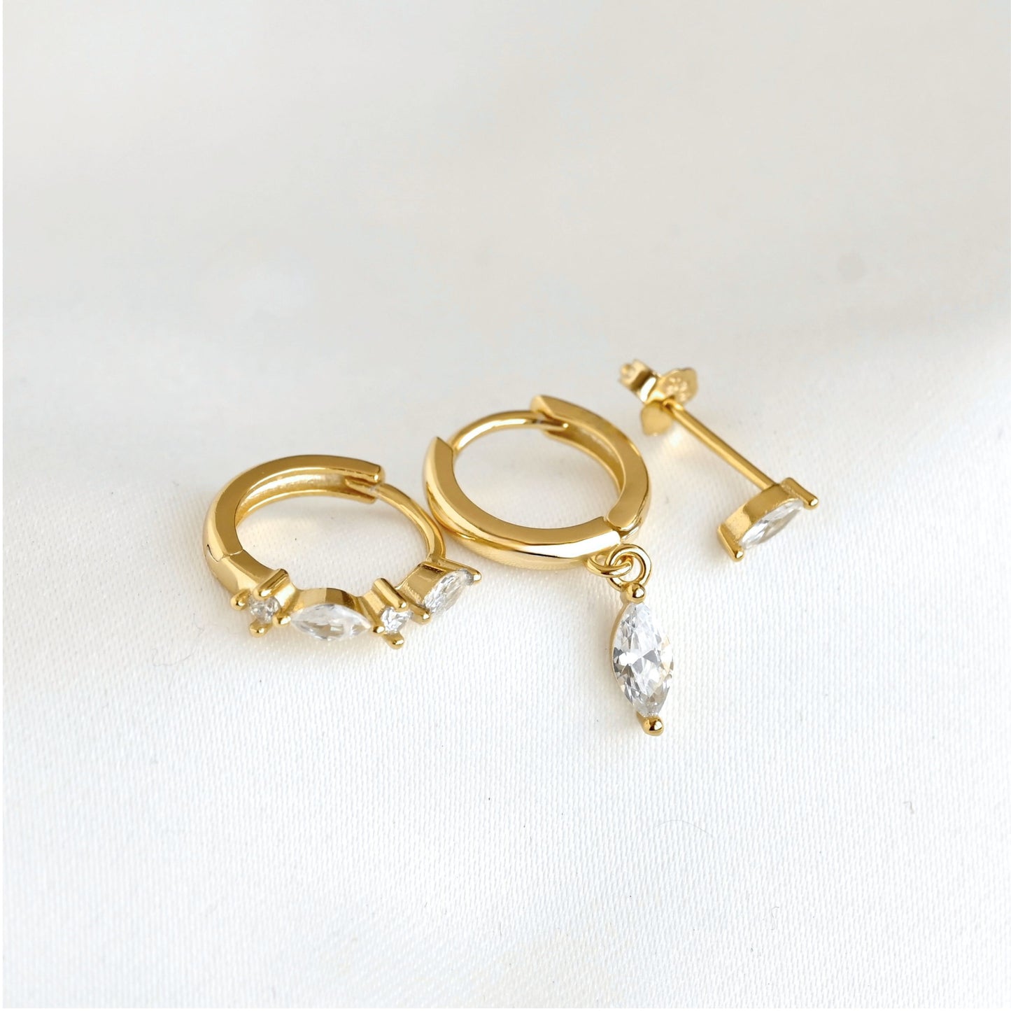 Gold huggie hoop earrings  and studs with marquise-cut clear crystals, shown on a clean white textured background.