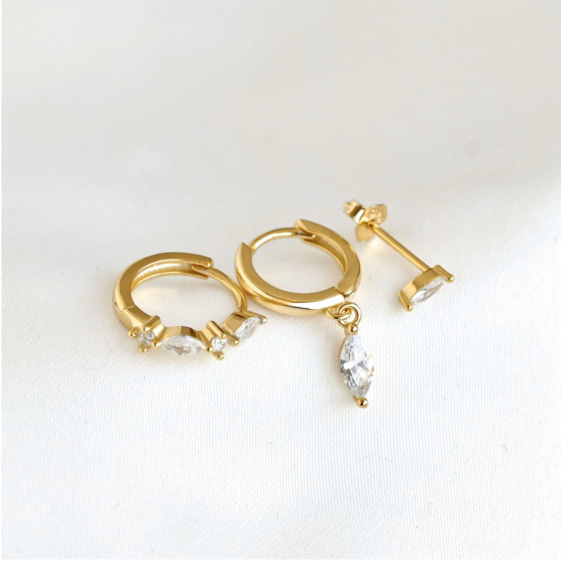 Gold huggie hoop earrings  and studs with marquise-cut clear crystals, shown on a clean white textured background.
