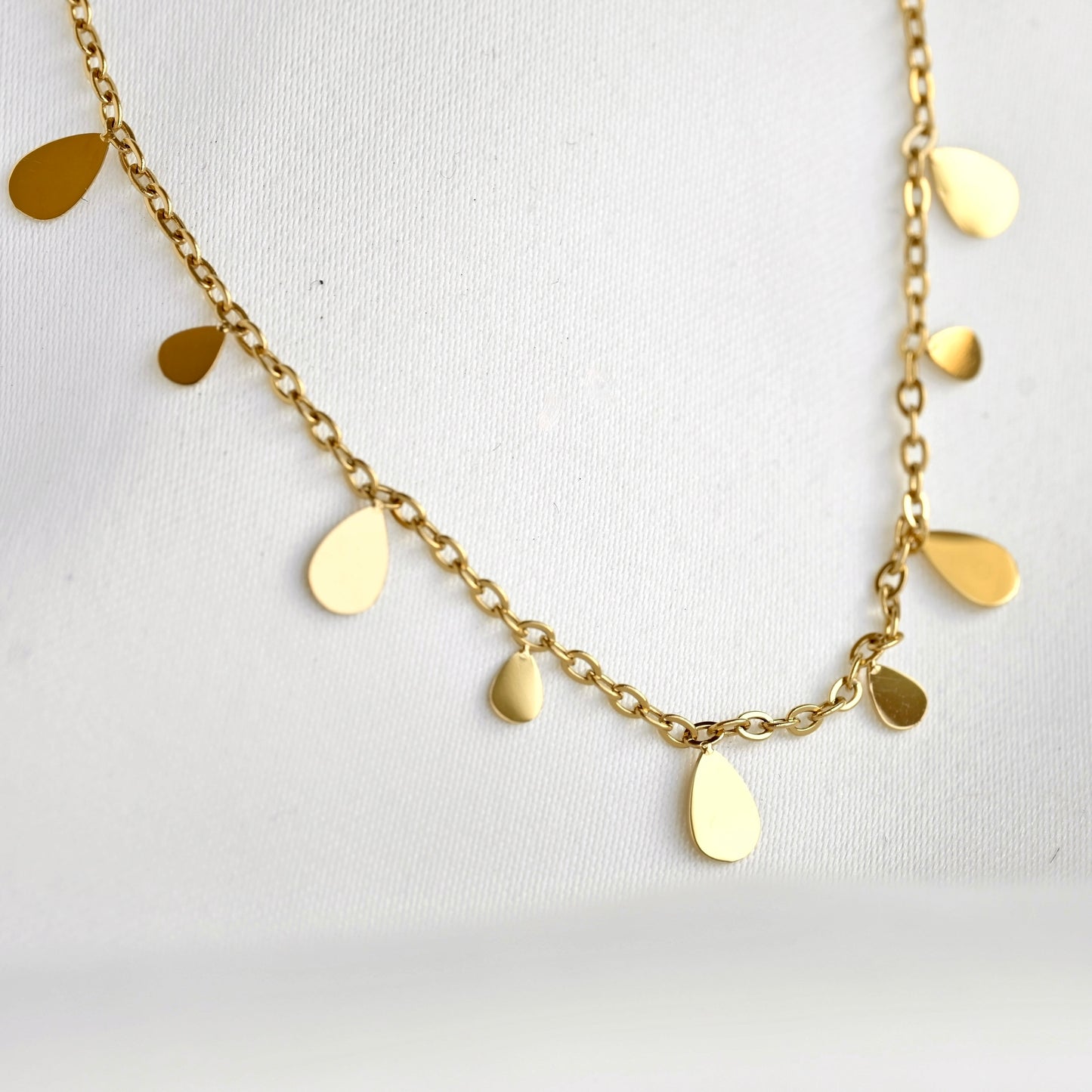 A gold chain necklace with small, irregular teardrop-shaped flat metal charms, displayed on a textured white fabric background.