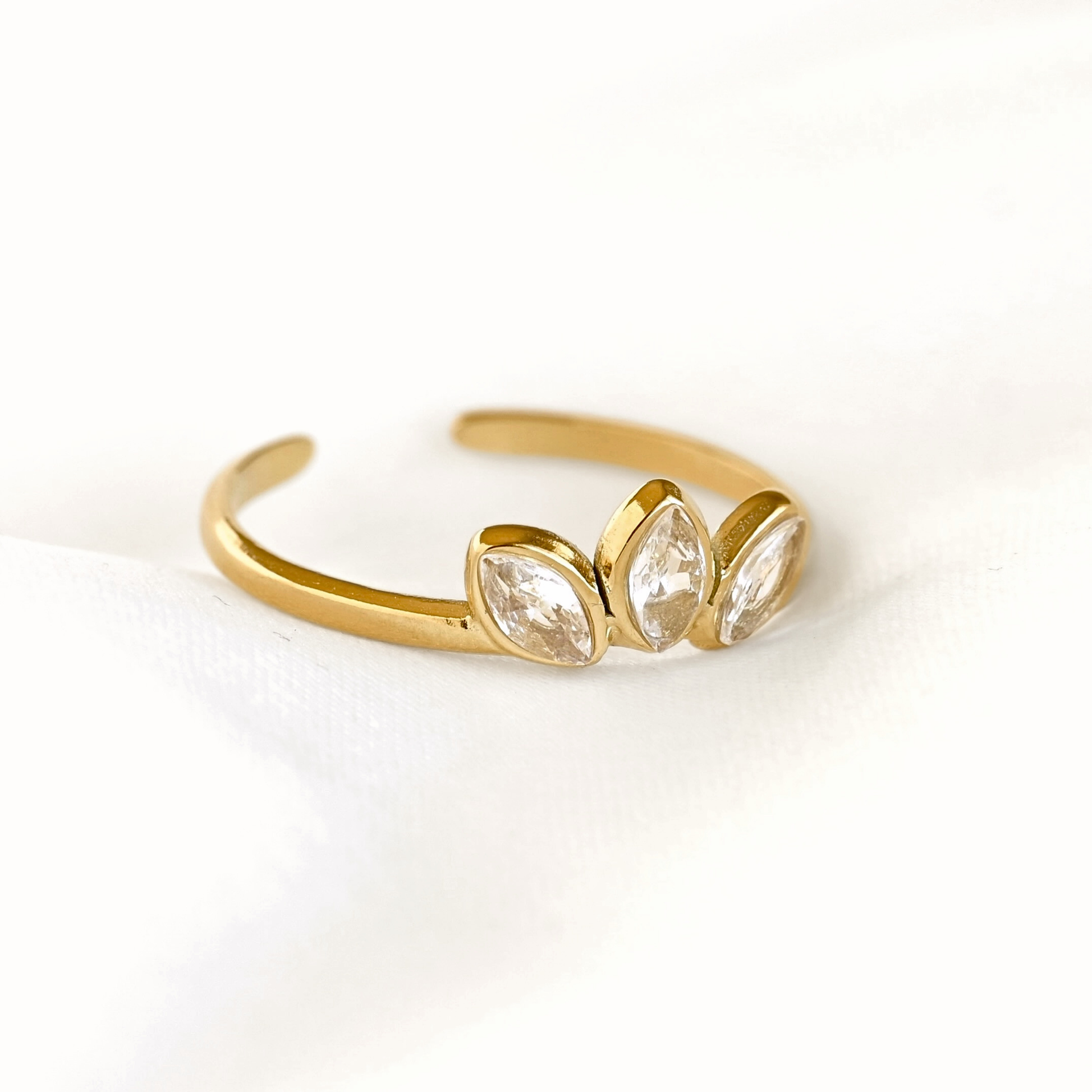 A gold adjustable ring featuring three marquise-cut clear crystals in a row, displayed on a soft white fabric background.