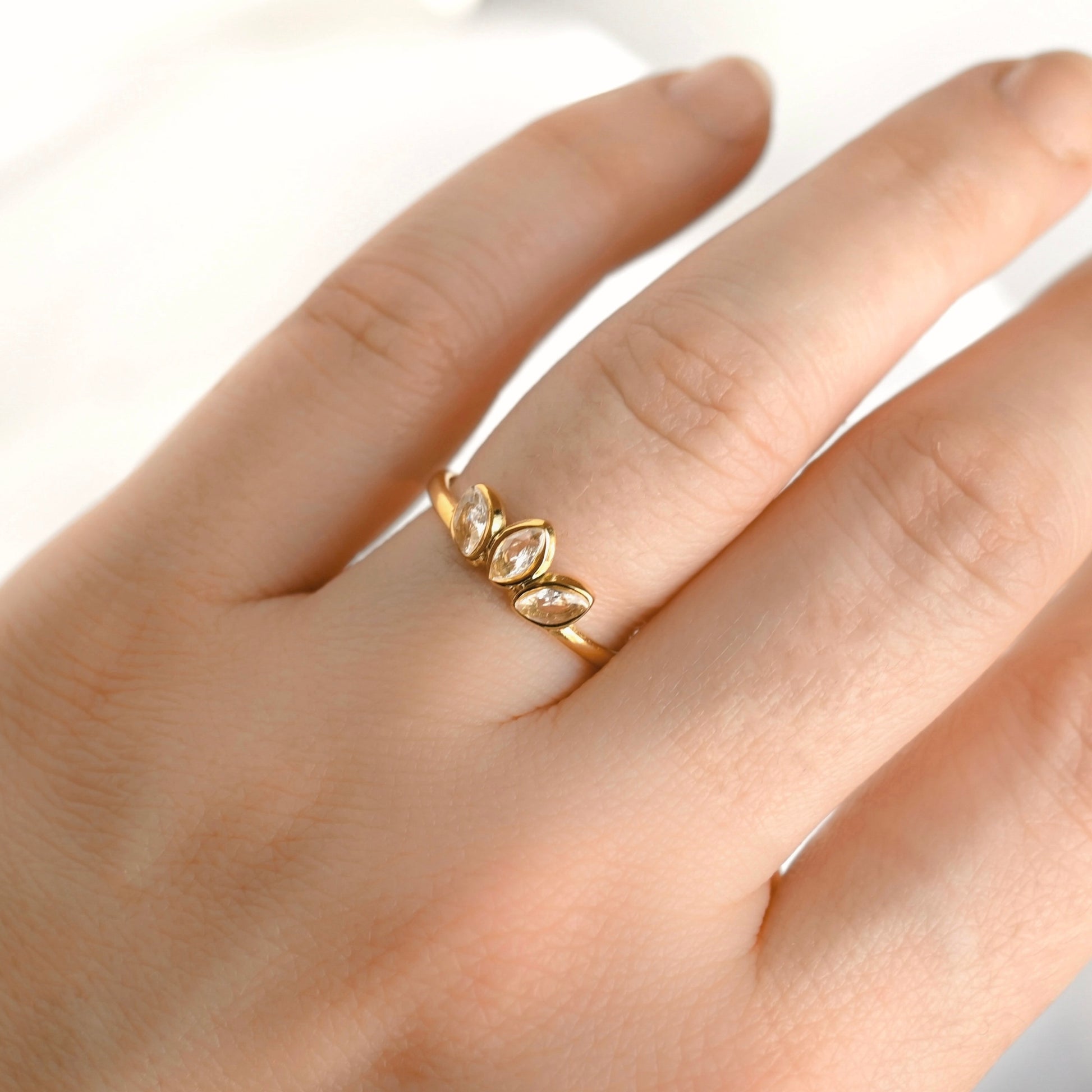 A close-up of a hand wearing a dainty gold ring with three marquise-cut clear stones set in a leaf-like cluster.
