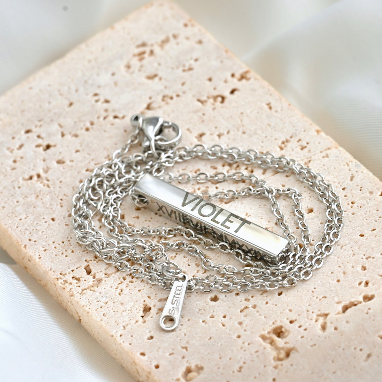 Silver cable chain necklace with a bar pendant engraved 'VIOLET' on a textured beige surface.