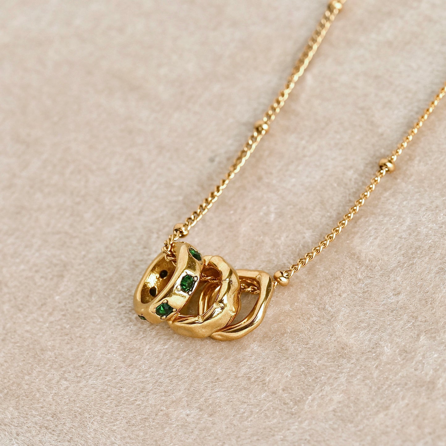 A gold necklace with three organic-shaped rings, displayed on a textured beige surface.