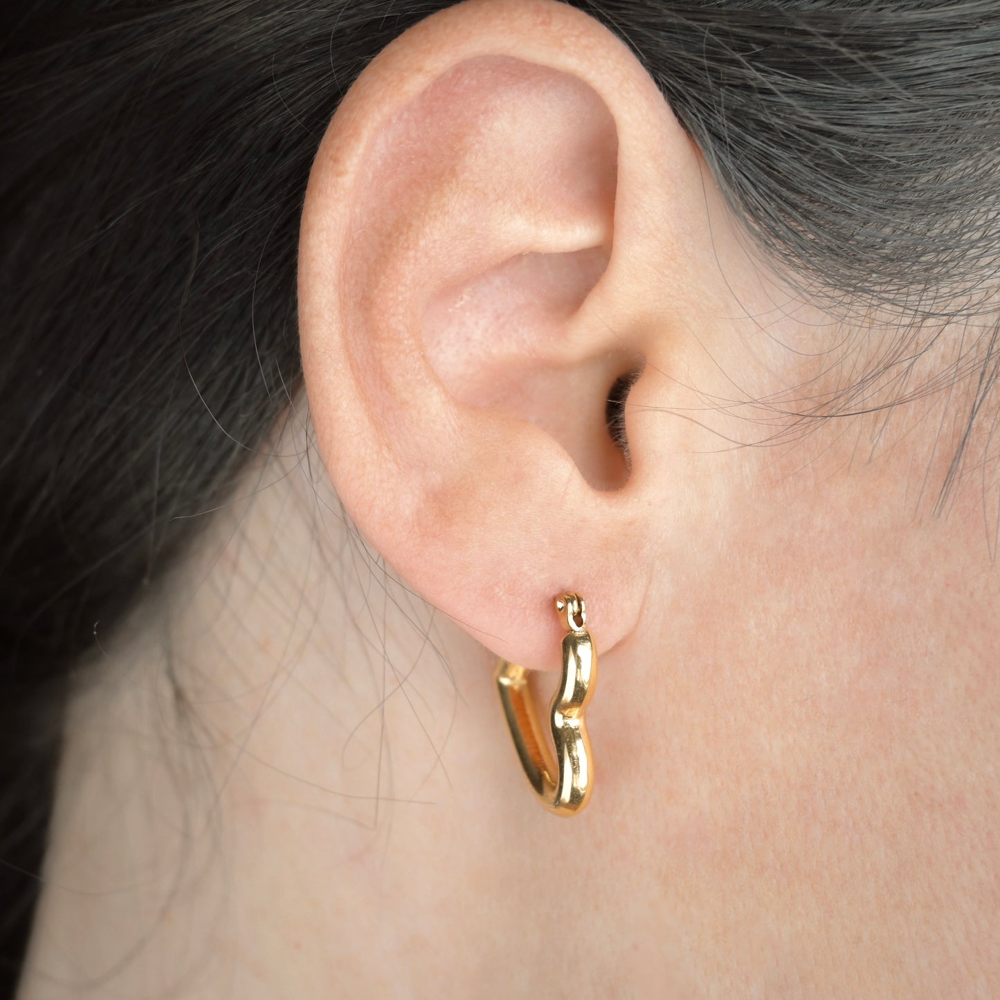Gold tube heart earring worn by a person with dark hair.