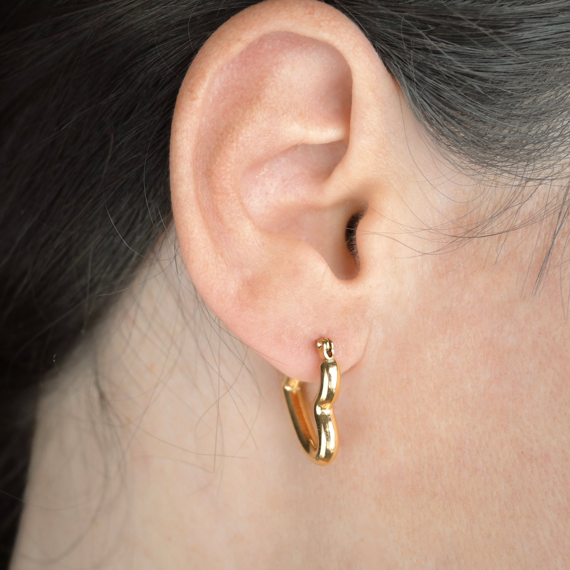 Gold tube heart earring worn by a person with dark hair.