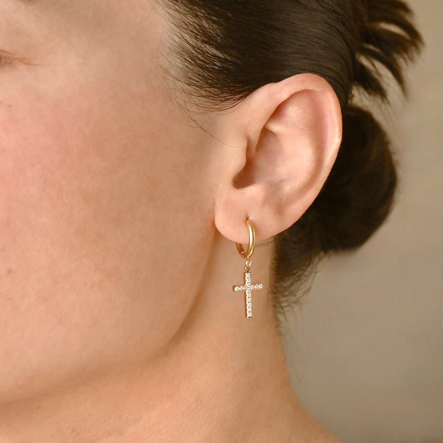 A close-up of a person's ear wearing  small gold hoop earrings with a dangling cross charm pave set with clear stones, against a soft beige background.