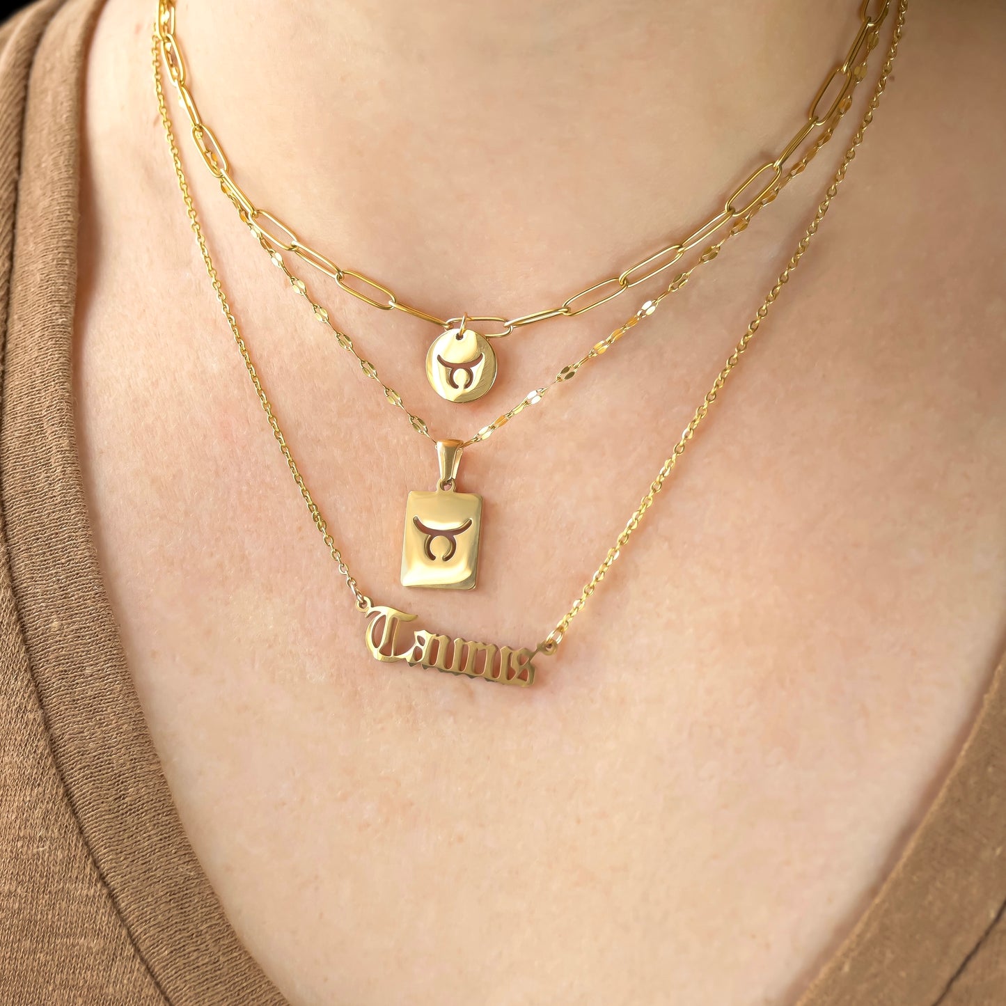 Front view of gold necklaces with taurus pendants on a person wearing a brown top: 1. a coin charm with taurus symbol cutout on a paperclip chain, 2. a tablet pendant with taurus symbol cutout  on a sequin chain, and 3. a spelled out Taurus script in gothic font on a cable chain.