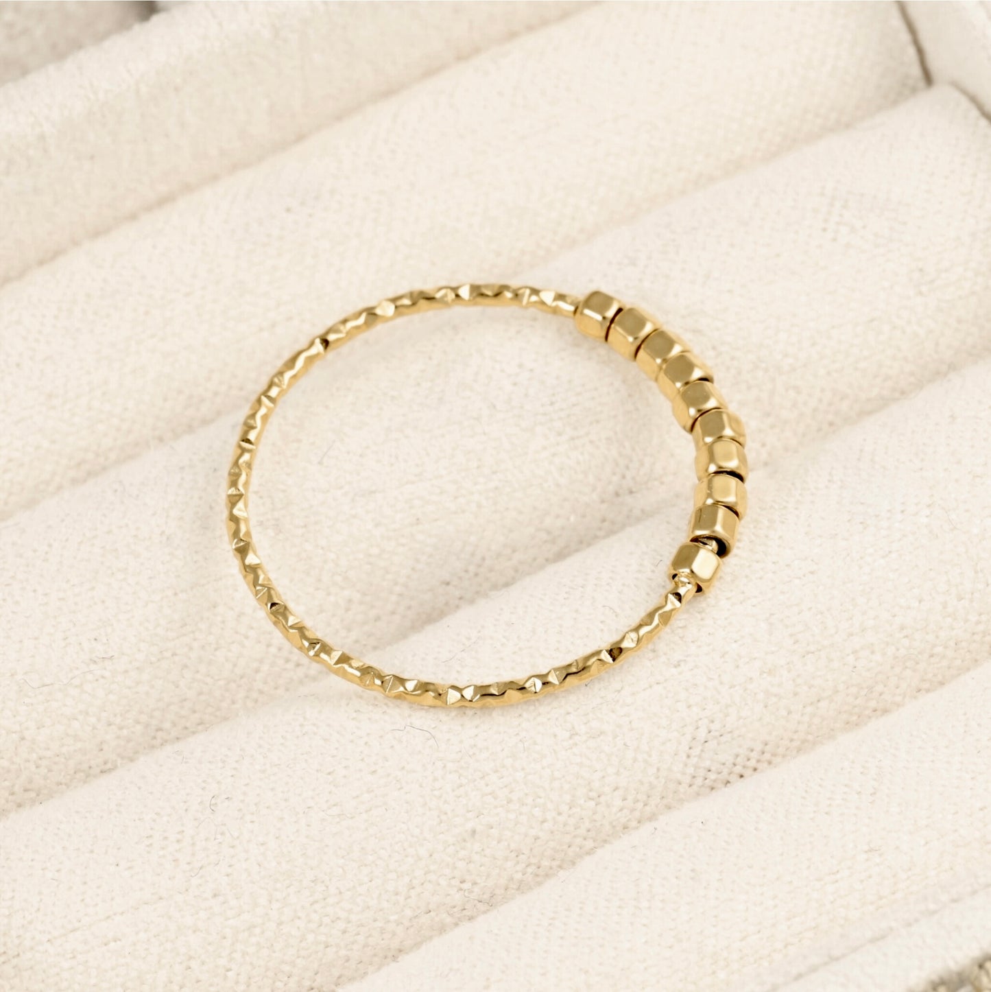 A gold beaded band ring with a textured, faceted bead pattern, displayed on a cream-colored fabric jewelry organizer.